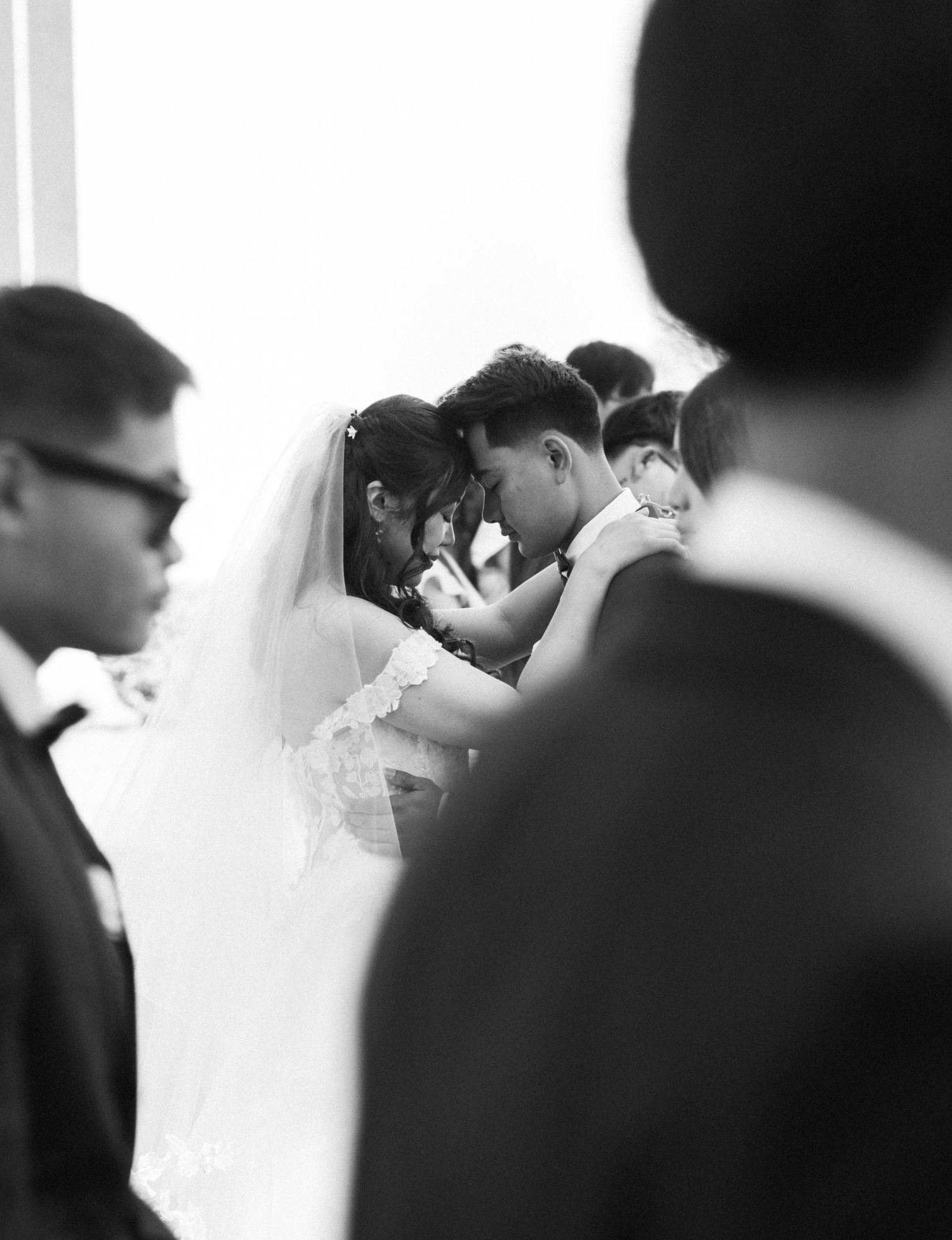 An intimate wedding moment captured in black and white with guests in the background.