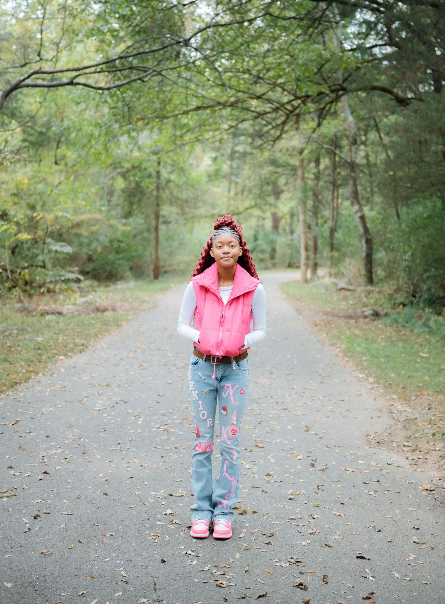 Person in pink vest and decorated jeans standing on a tree-lined path.