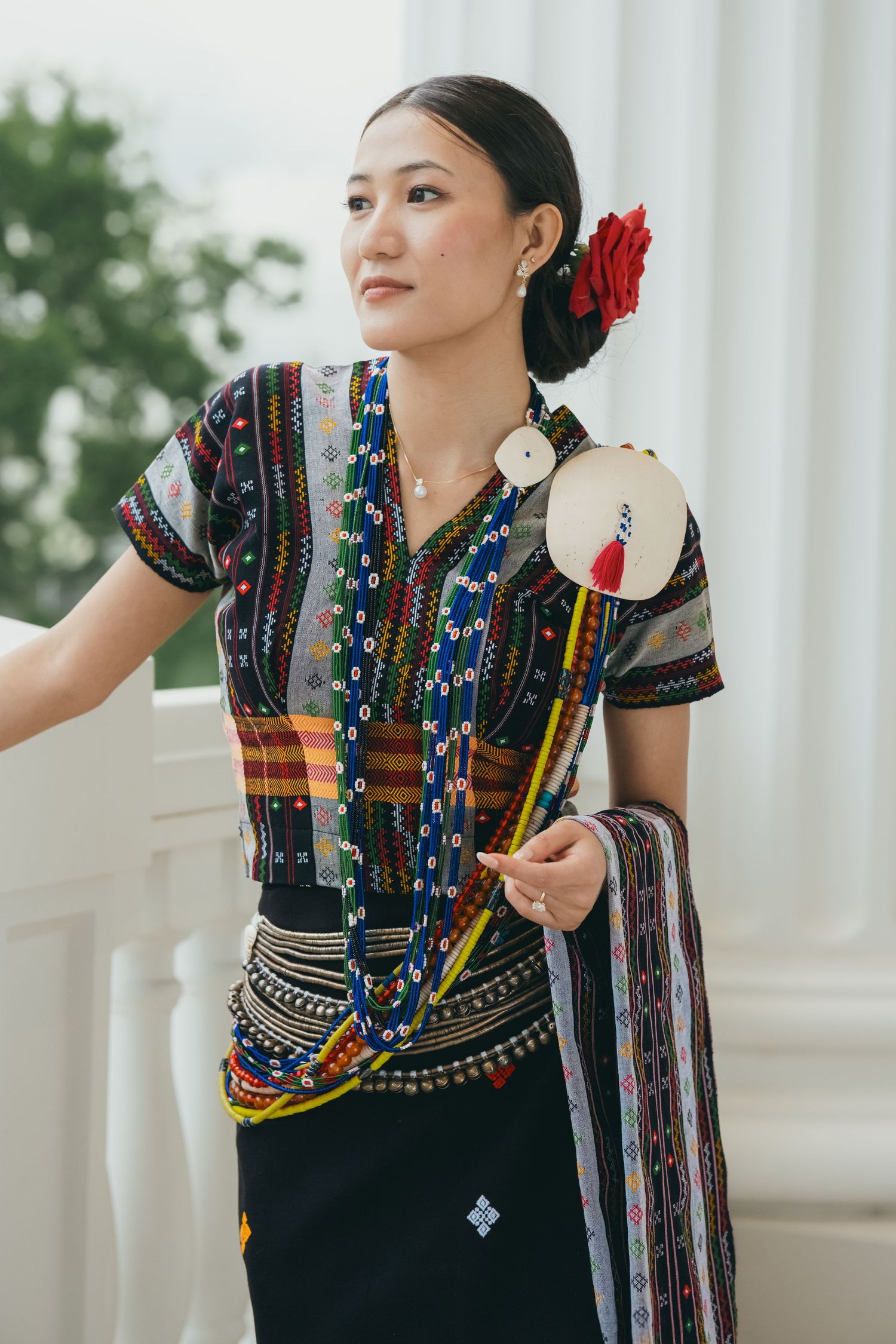 Ornate traditional dress with beaded necklaces and red flower accessory photographed by columns.