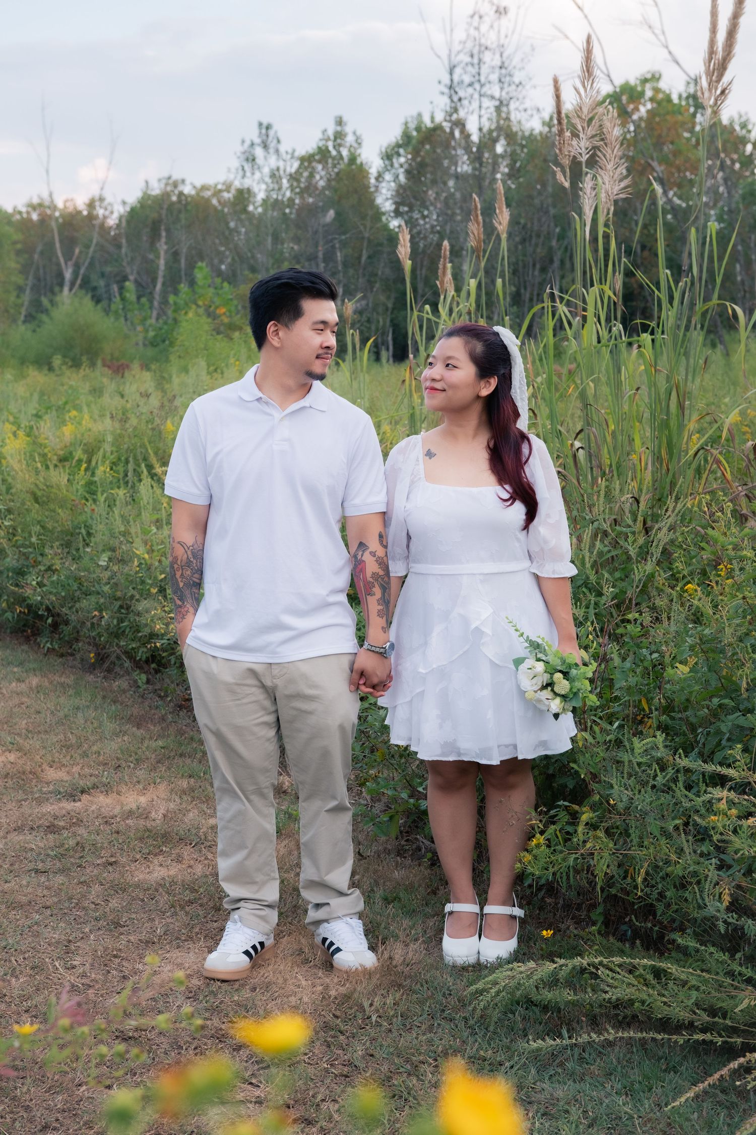 A couple holds hands while walking on a nature trail surrounded by tall grass and wildflowers.