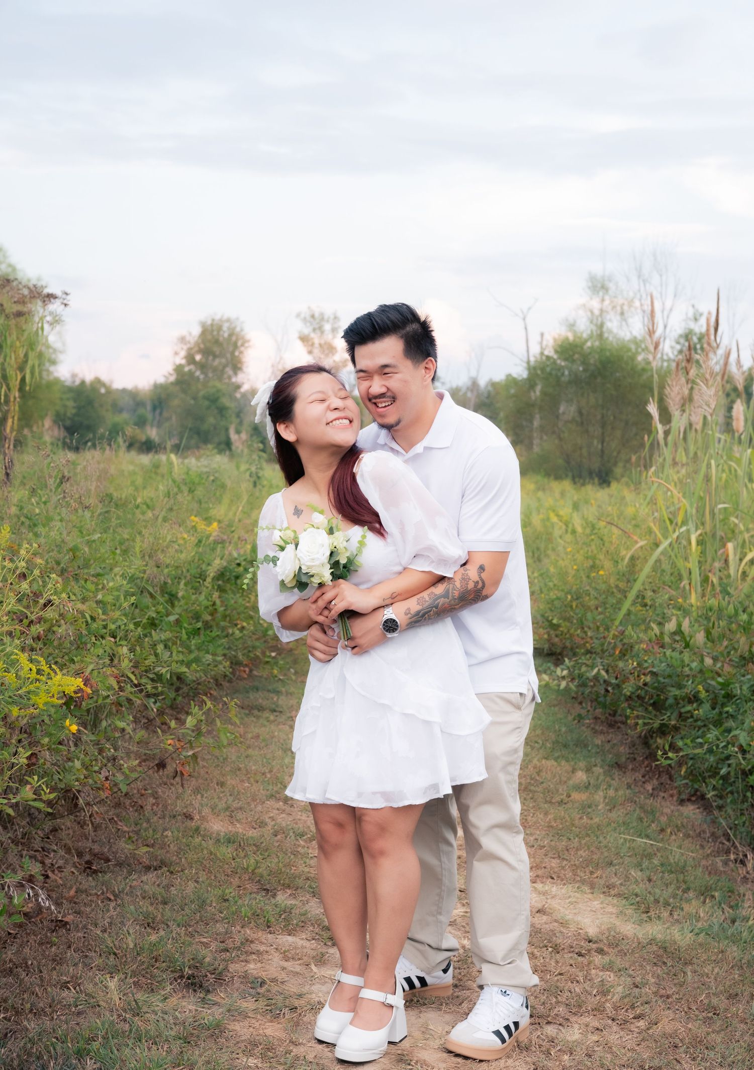A couple dressed in white shares a romantic moment on a nature path surrounded by greenery.