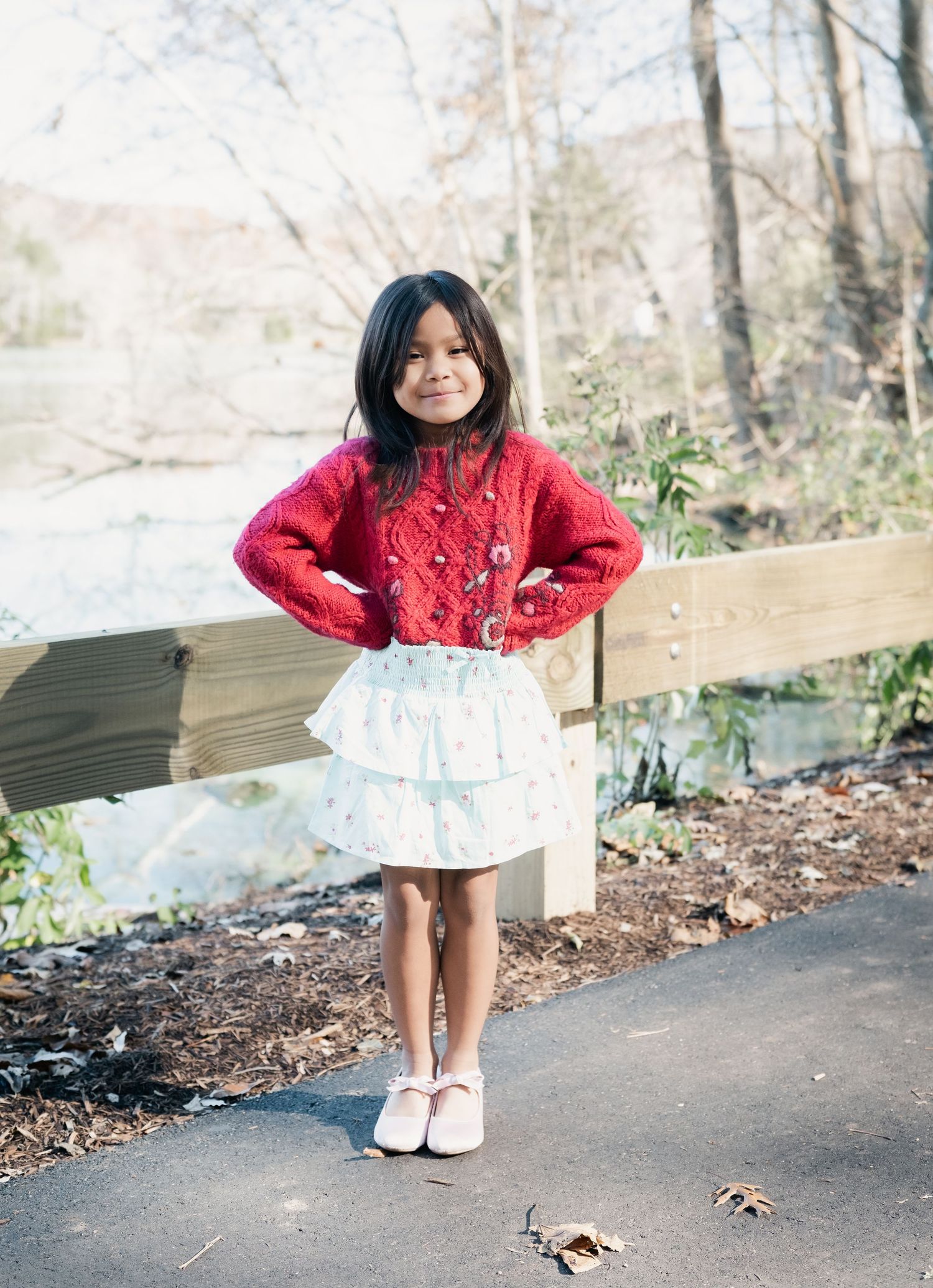 Child in red sweater and white dress posing playfully on outdoor path.