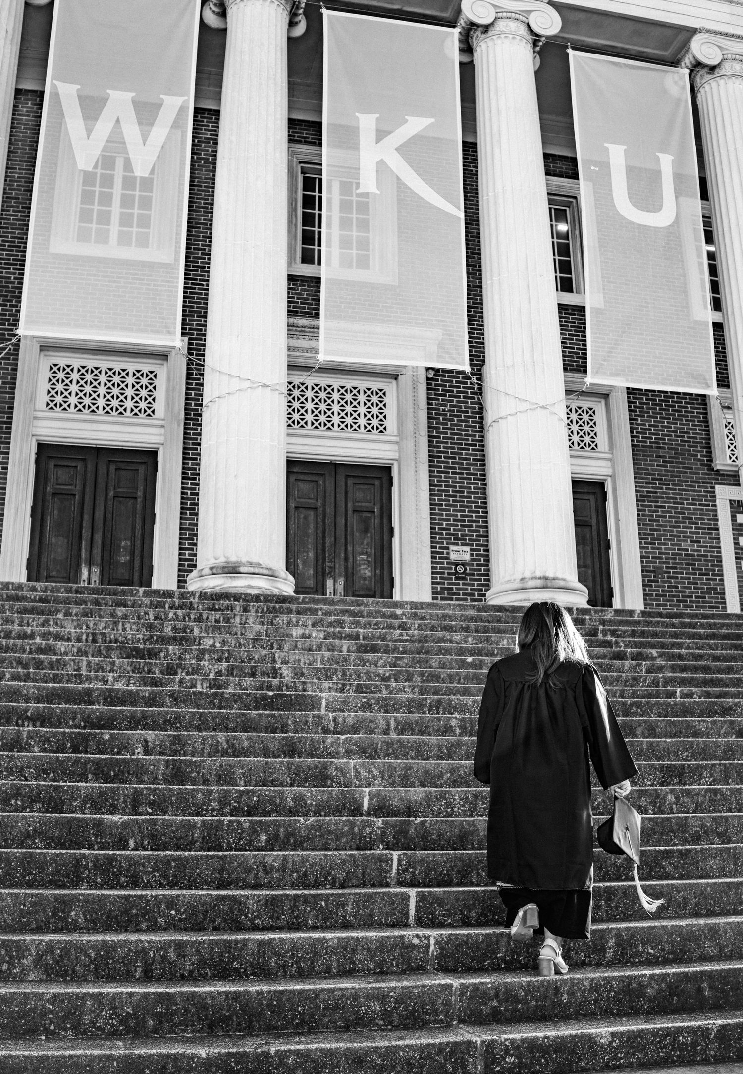 Graduate climbing steps at WKU campus with large white banners in monochrome.