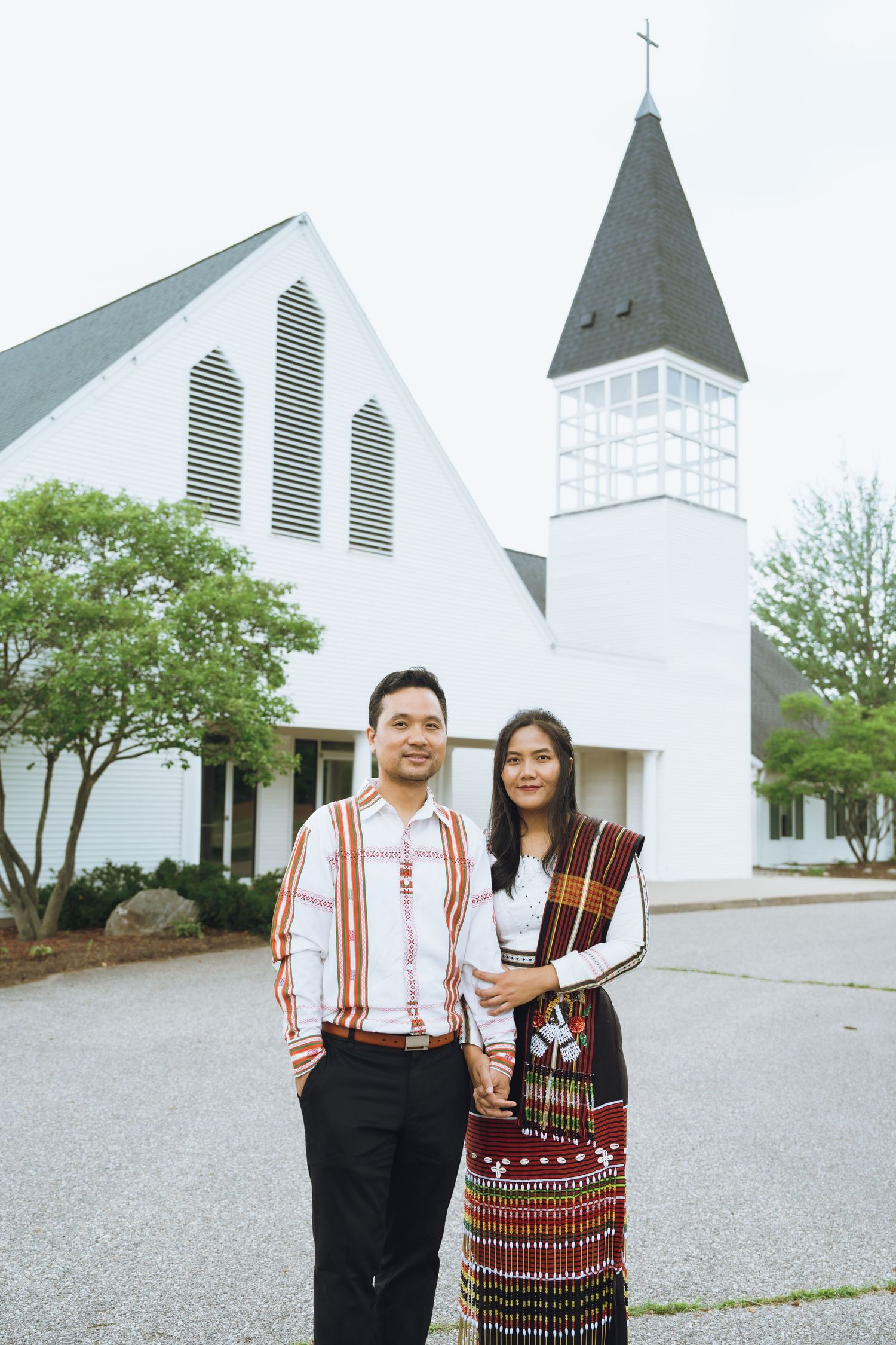 Couple in traditional Karen attire posing in front of a white church with a steeple on a cloudy day.