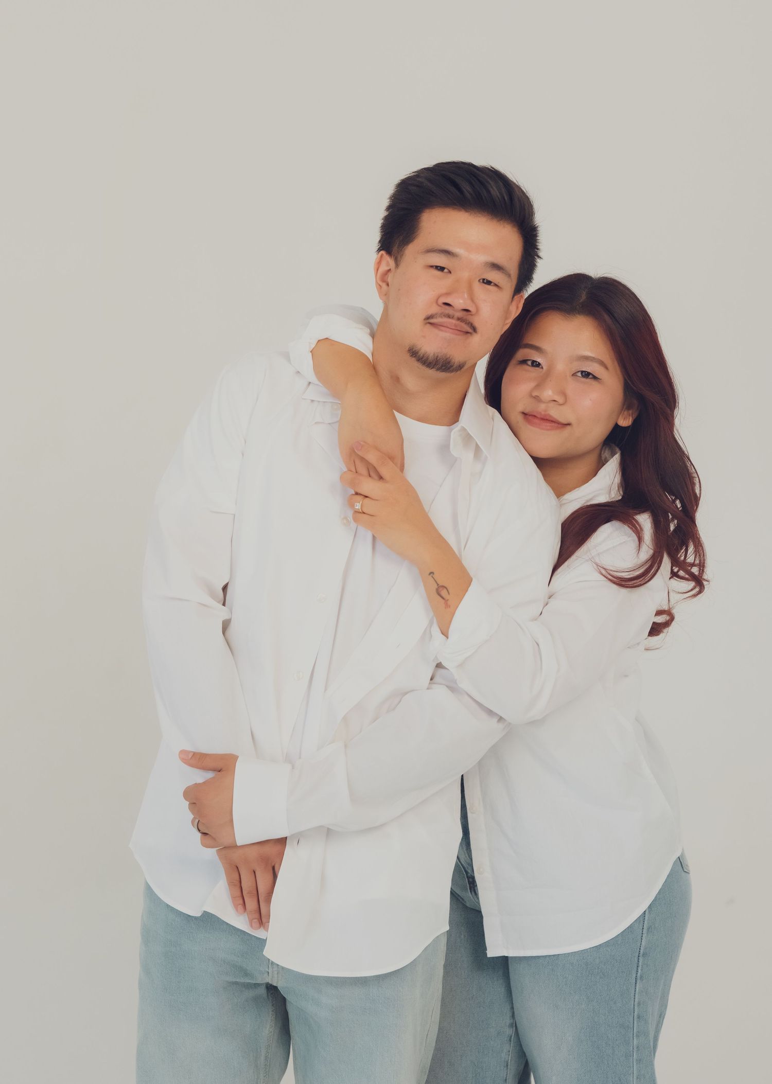 A couple in matching white shirts and jeans pose together, the woman hugging the man from behind against a light background.