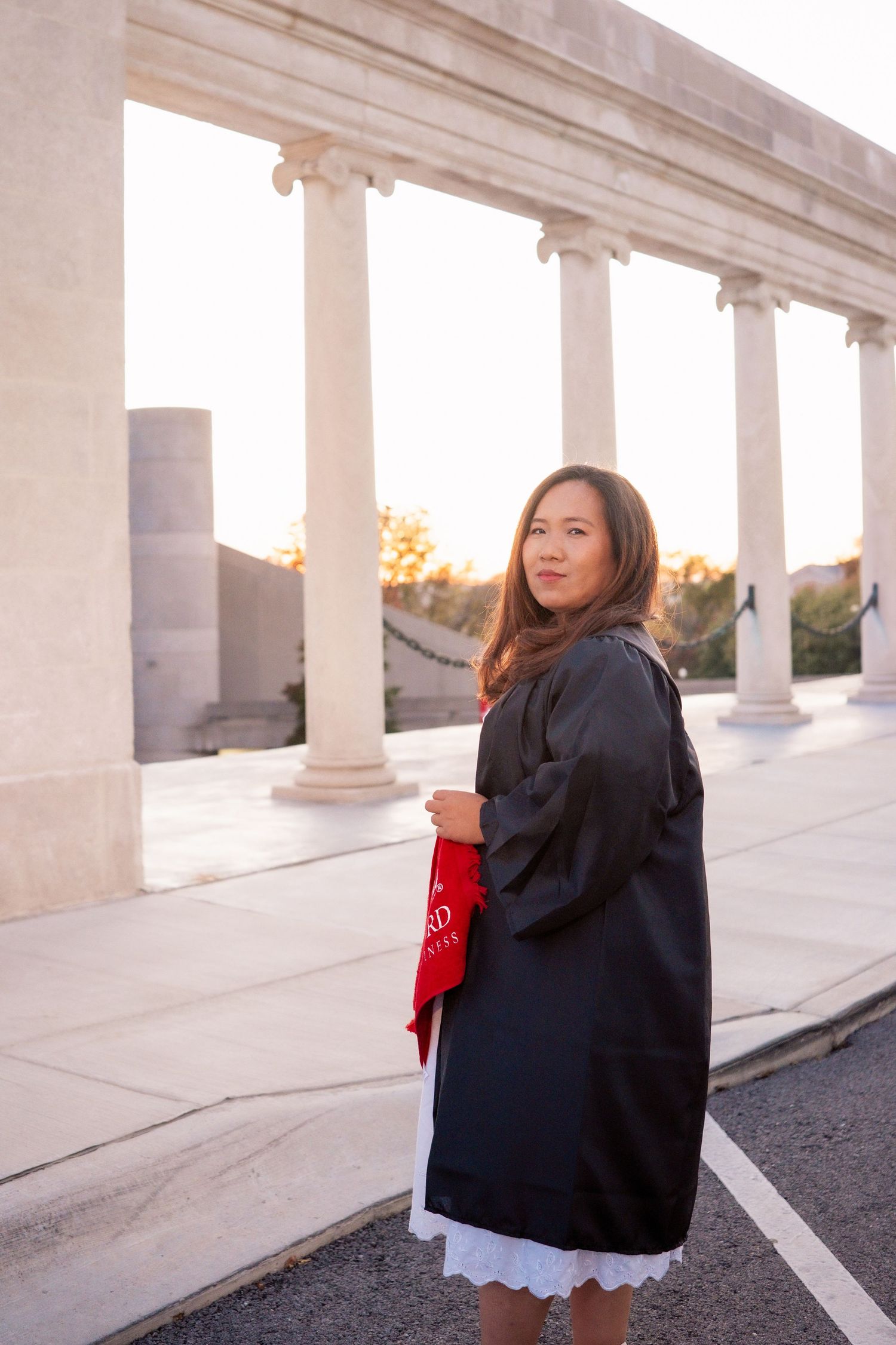 Graduate in black cap and gown poses along white columned walkway during sunset.
