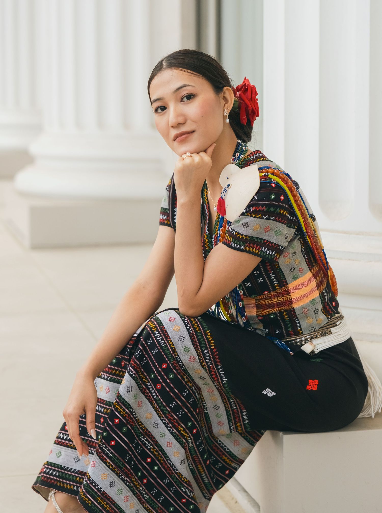 Person in traditional patterned dress poses elegantly against white columns.