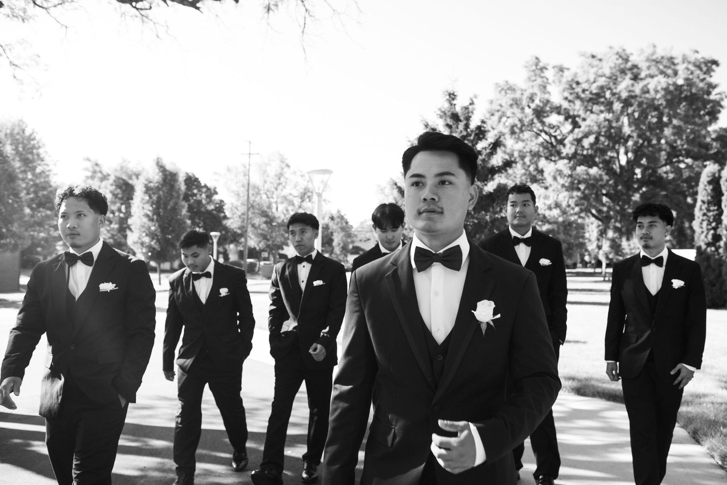 Groomsmen in black tuxedos walk in a line outdoors.