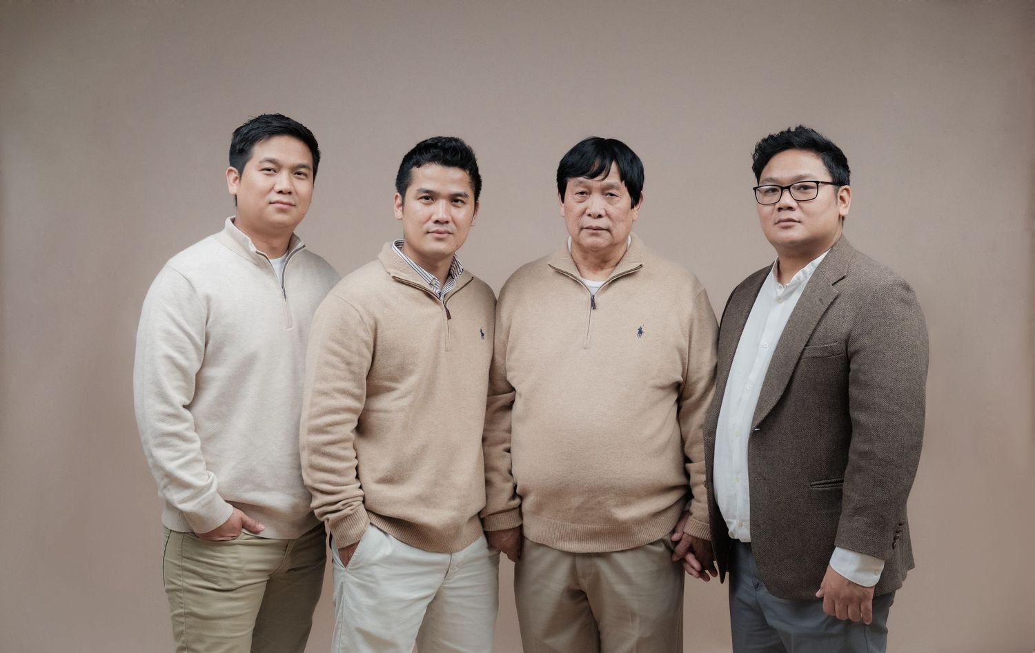 Four people in coordinated neutral-toned outfits pose against a brown backdrop for a professional portrait.