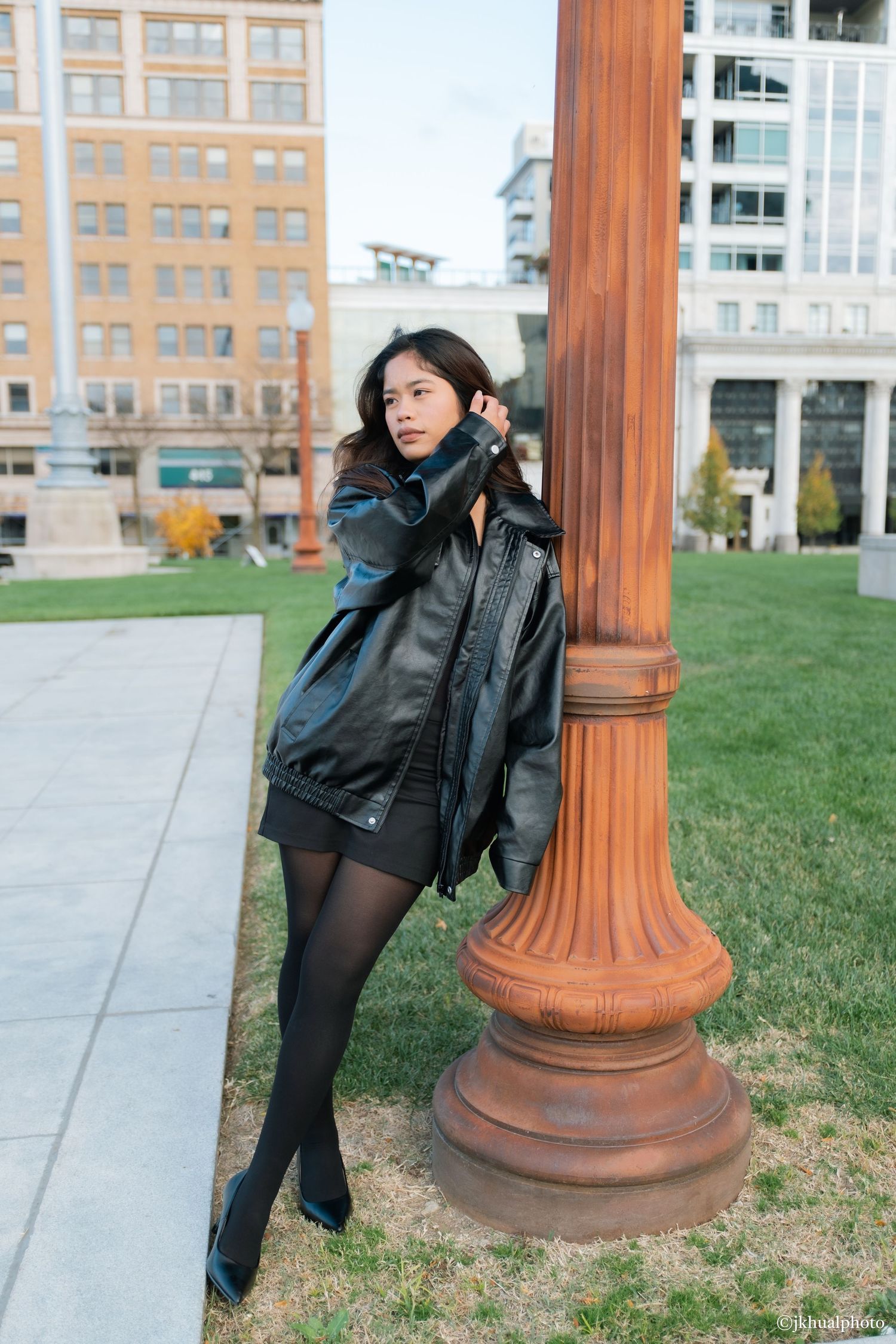 A person in a black leather jacket poses playfully by a decorative terracotta column on a grassy lawn near buildings.