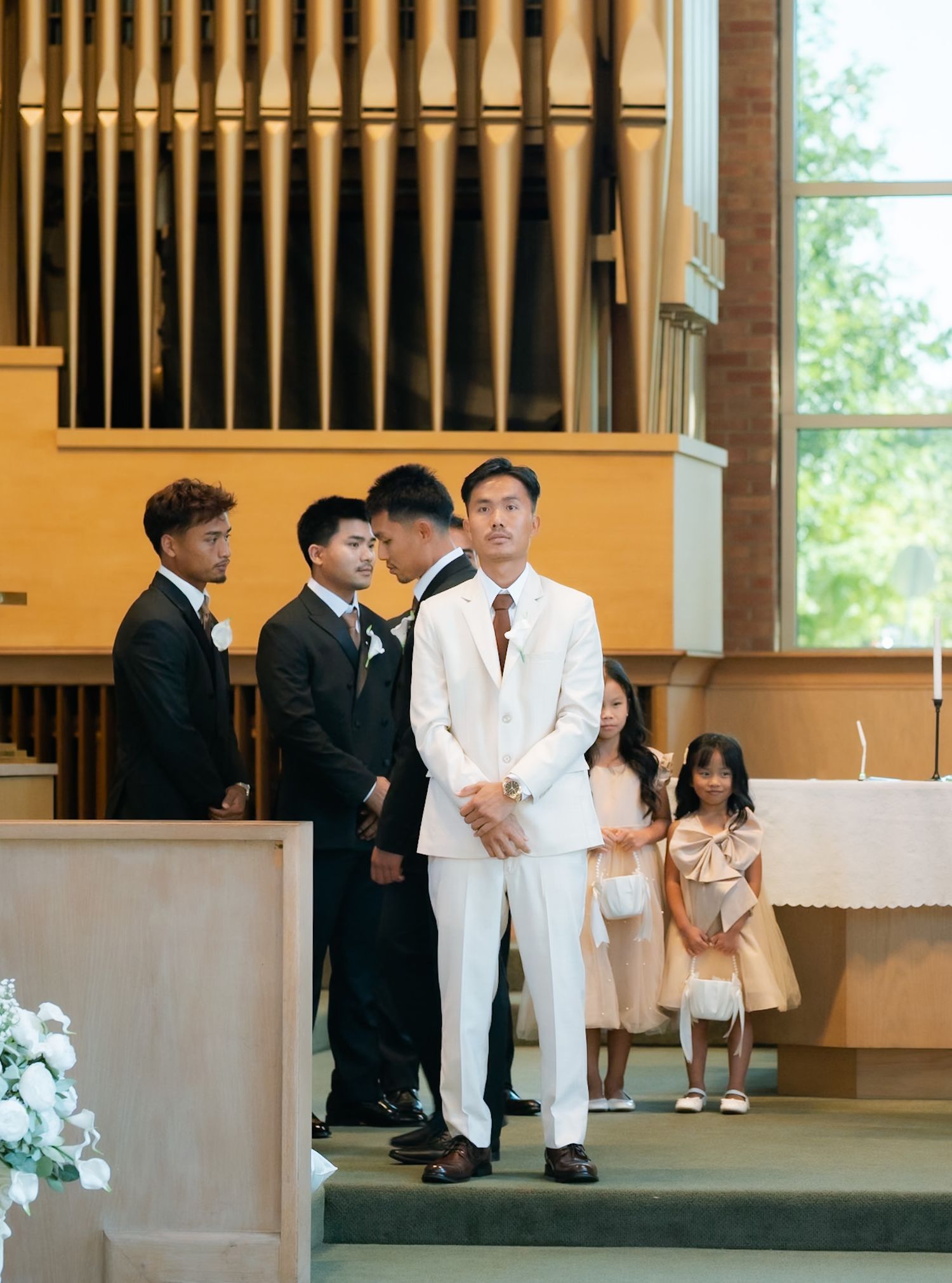 Groom in white suit stands at church altar with groomsmen in black suits and flower girls during wedding ceremony.