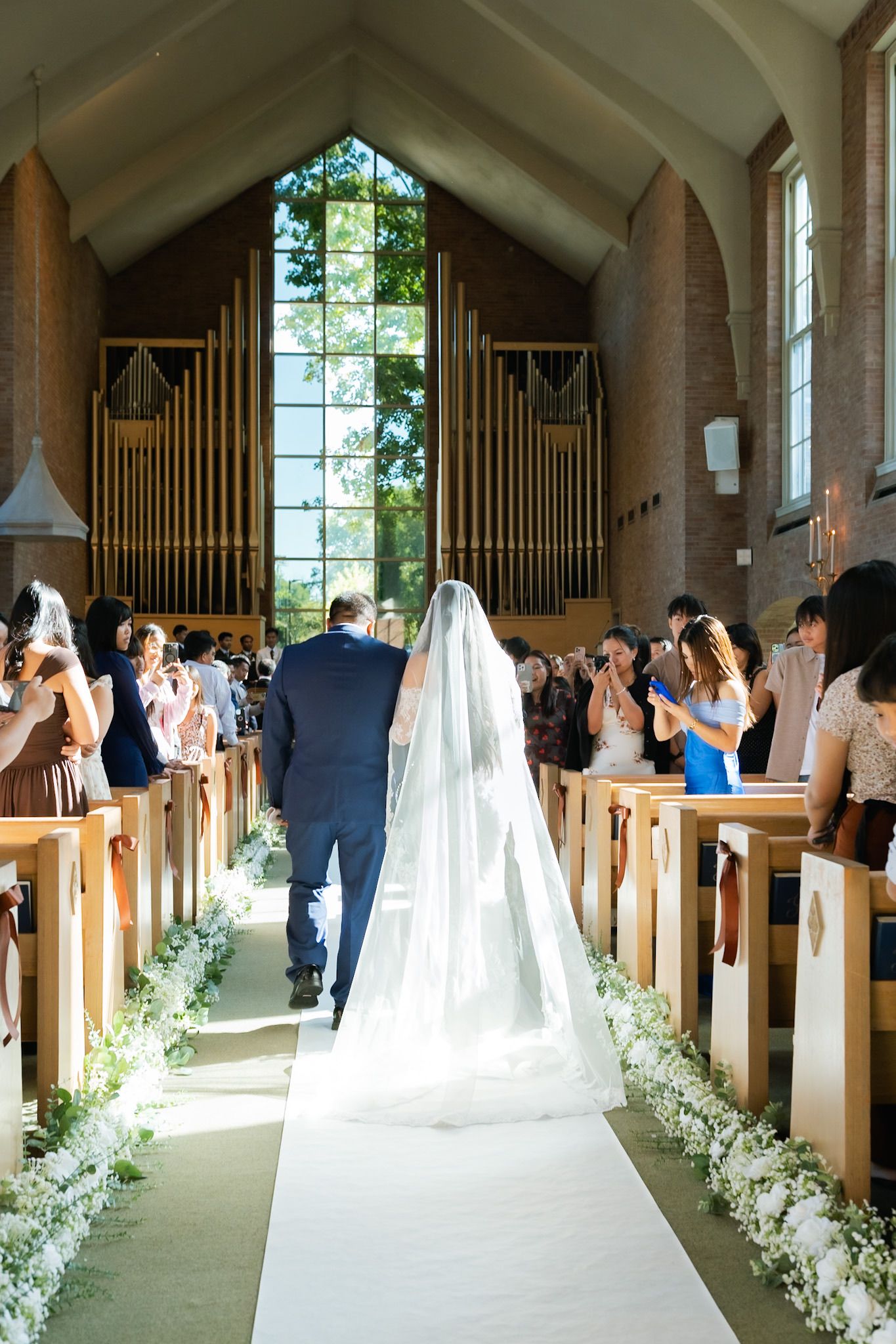 Bride walking down church aisle with escort, floral arrangements lining white runner, pipe organ and stained glass in background.