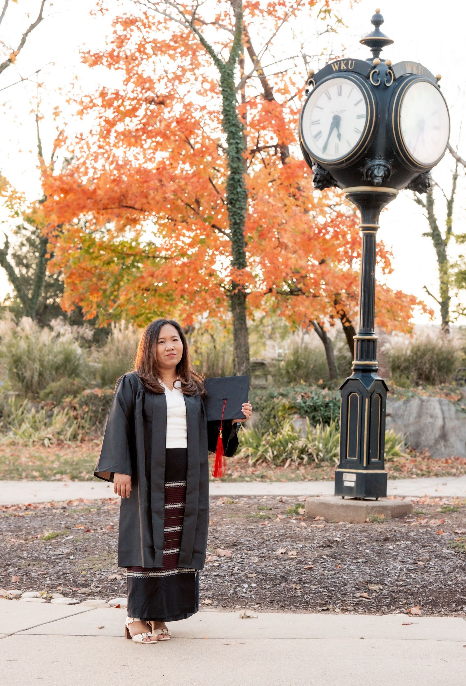 Graduate poses near decorative clock post with autumn foliage in background.