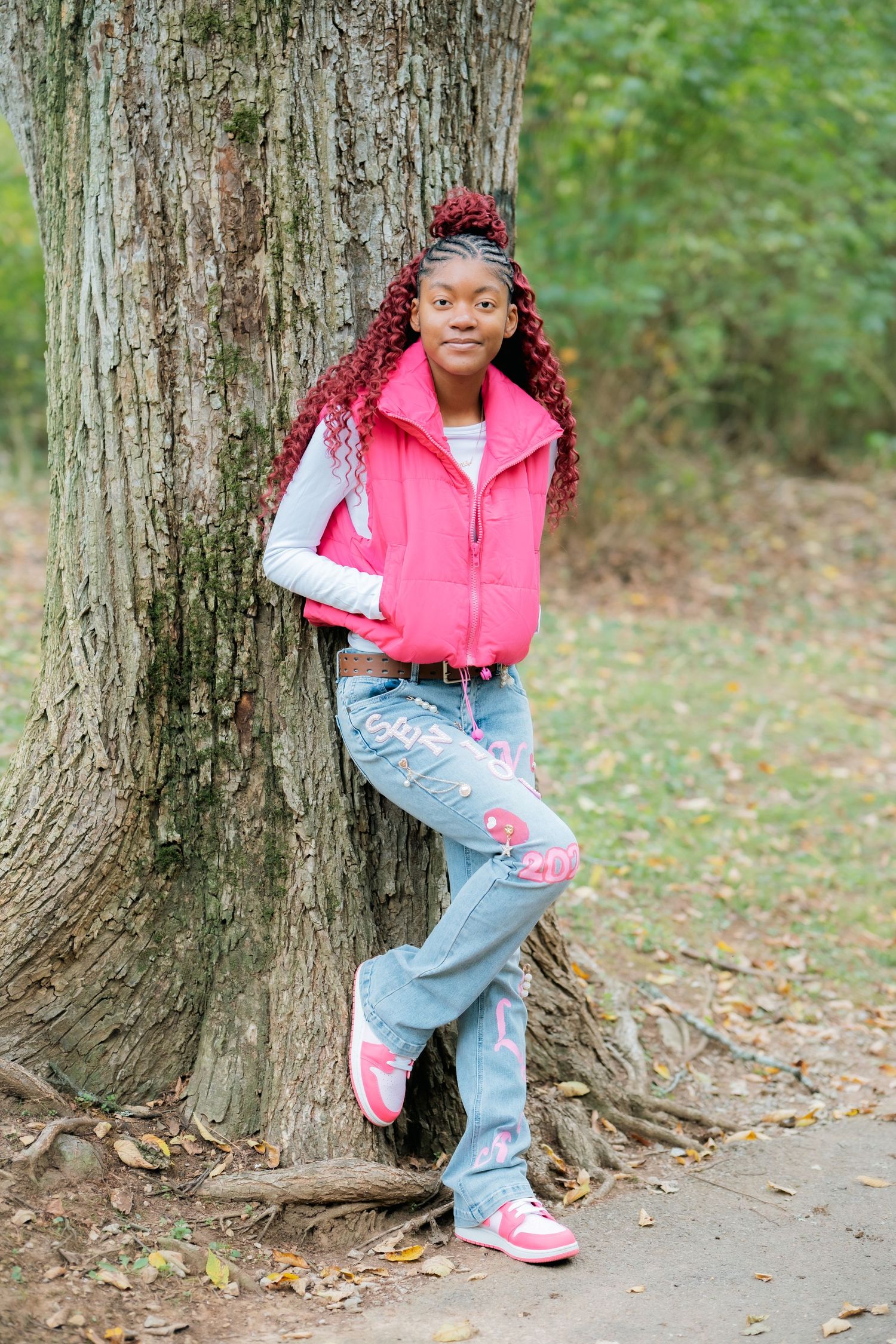 Stylish pose against large tree trunk wearing pink vest and decorated jeans.