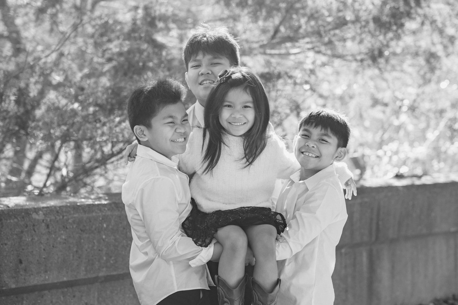 A playful outdoor photoshoot showing three children in white shirts and dark pants posing together on a path.