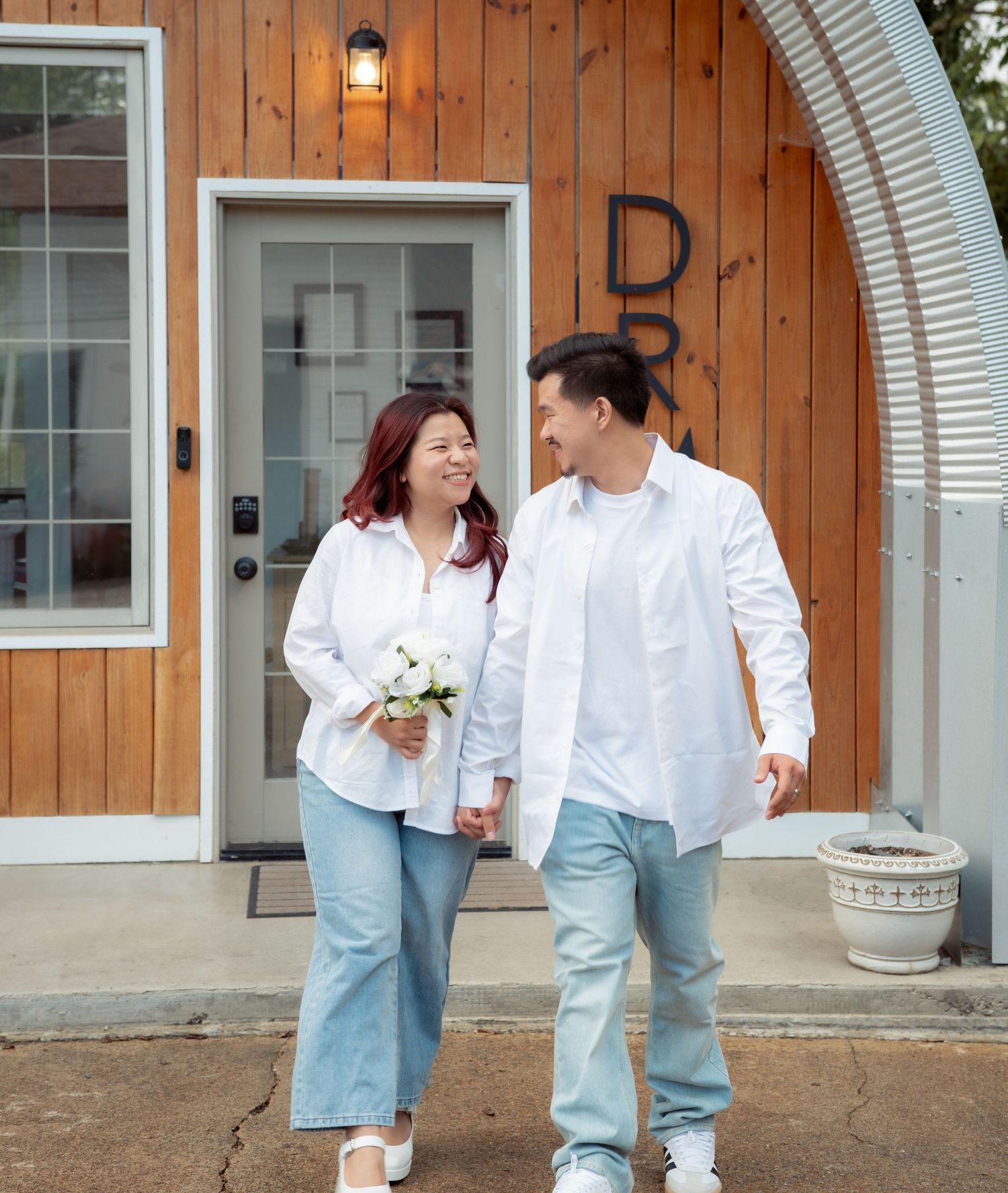 A couple in matching casual white and denim outfits poses together against wooden paneling.