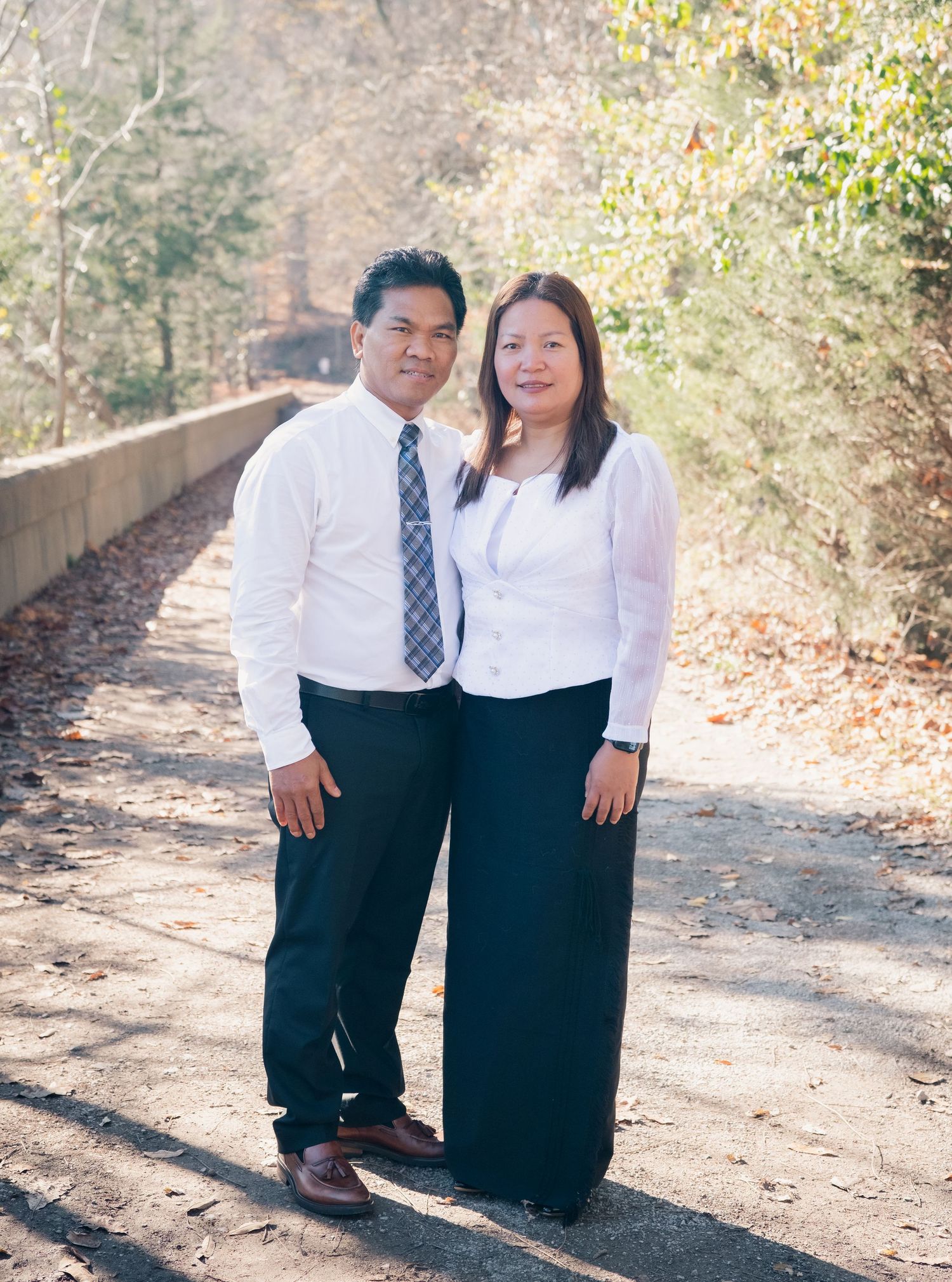 A couple stands close together on a leaf-strewn path wearing coordinated formal attire.