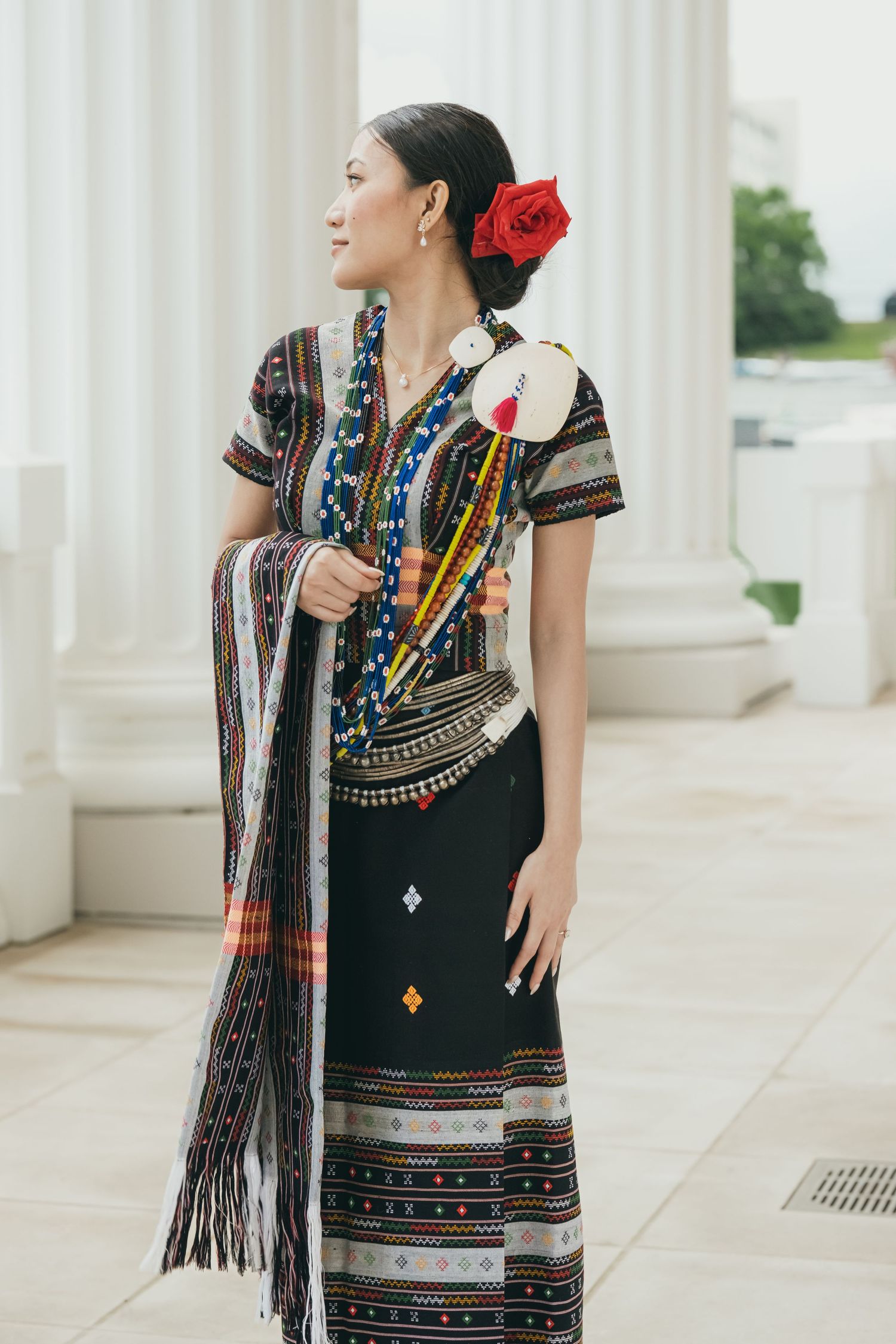 Traditional ethnic costume with colorful embroidery and red flower accessory photographed by white columns.