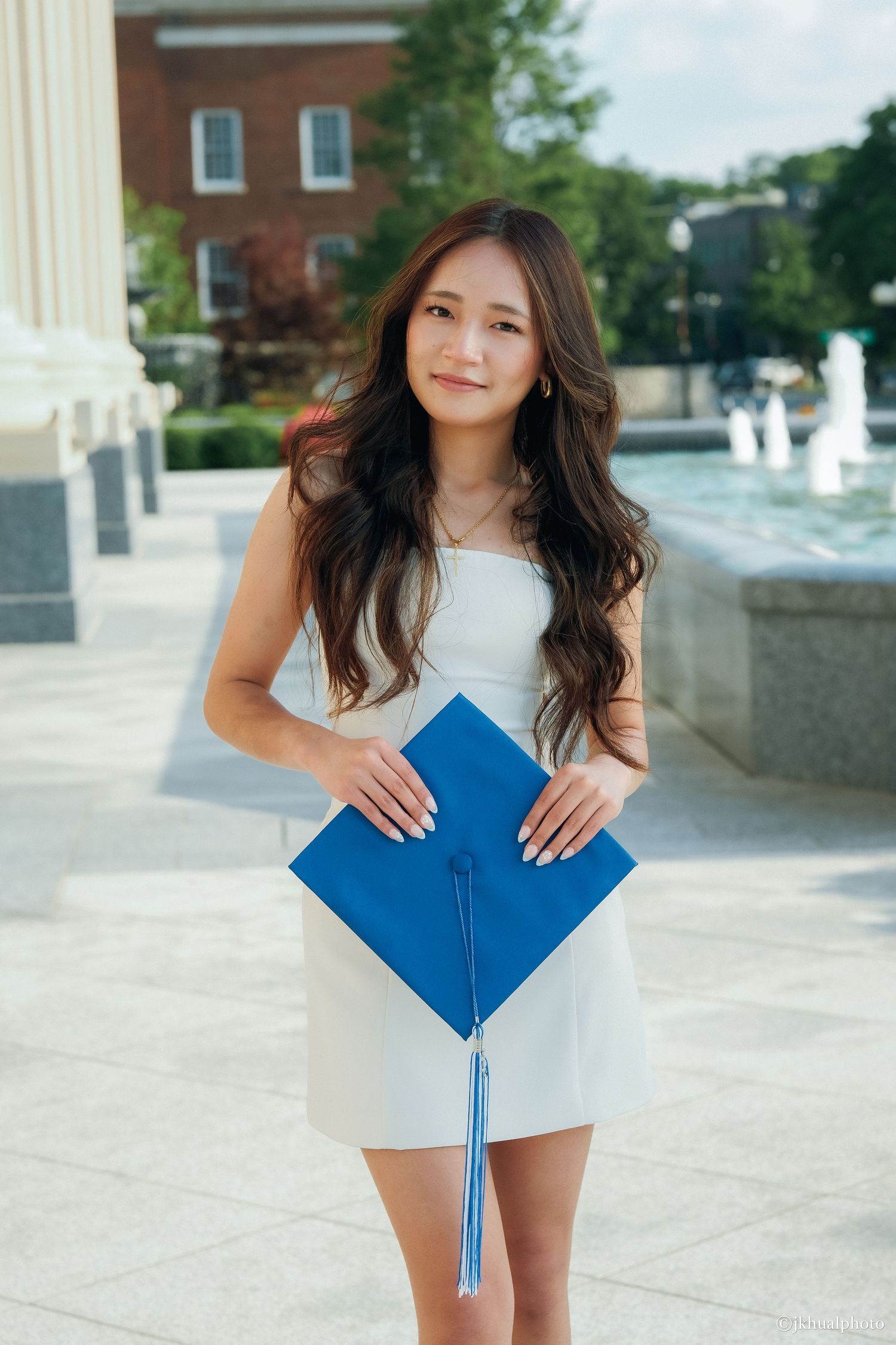 Graduate in white dress poses with blue diploma on sunny campus.