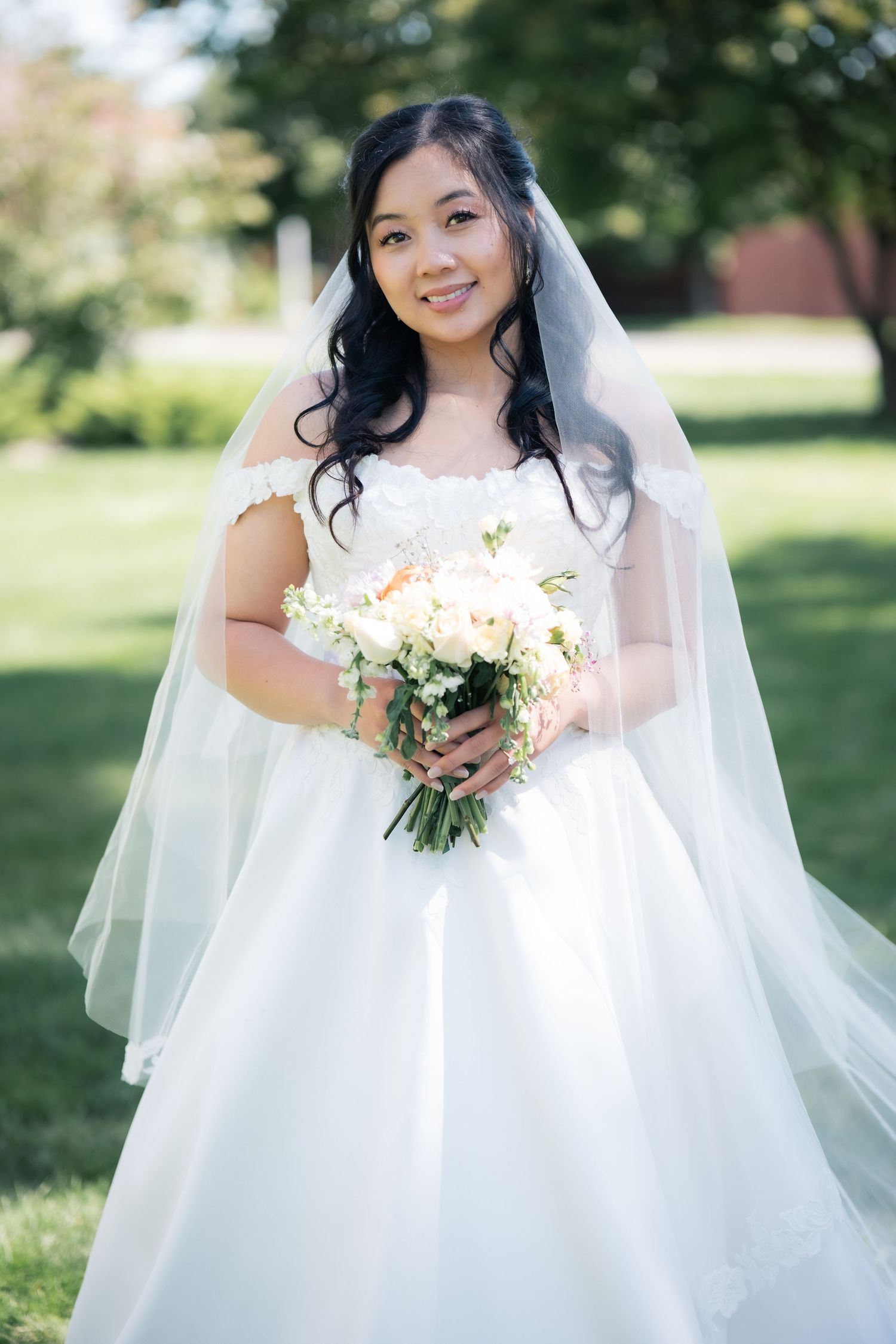 Radiant bride in off-shoulder wedding gown poses with white floral bouquet on sunny lawn.