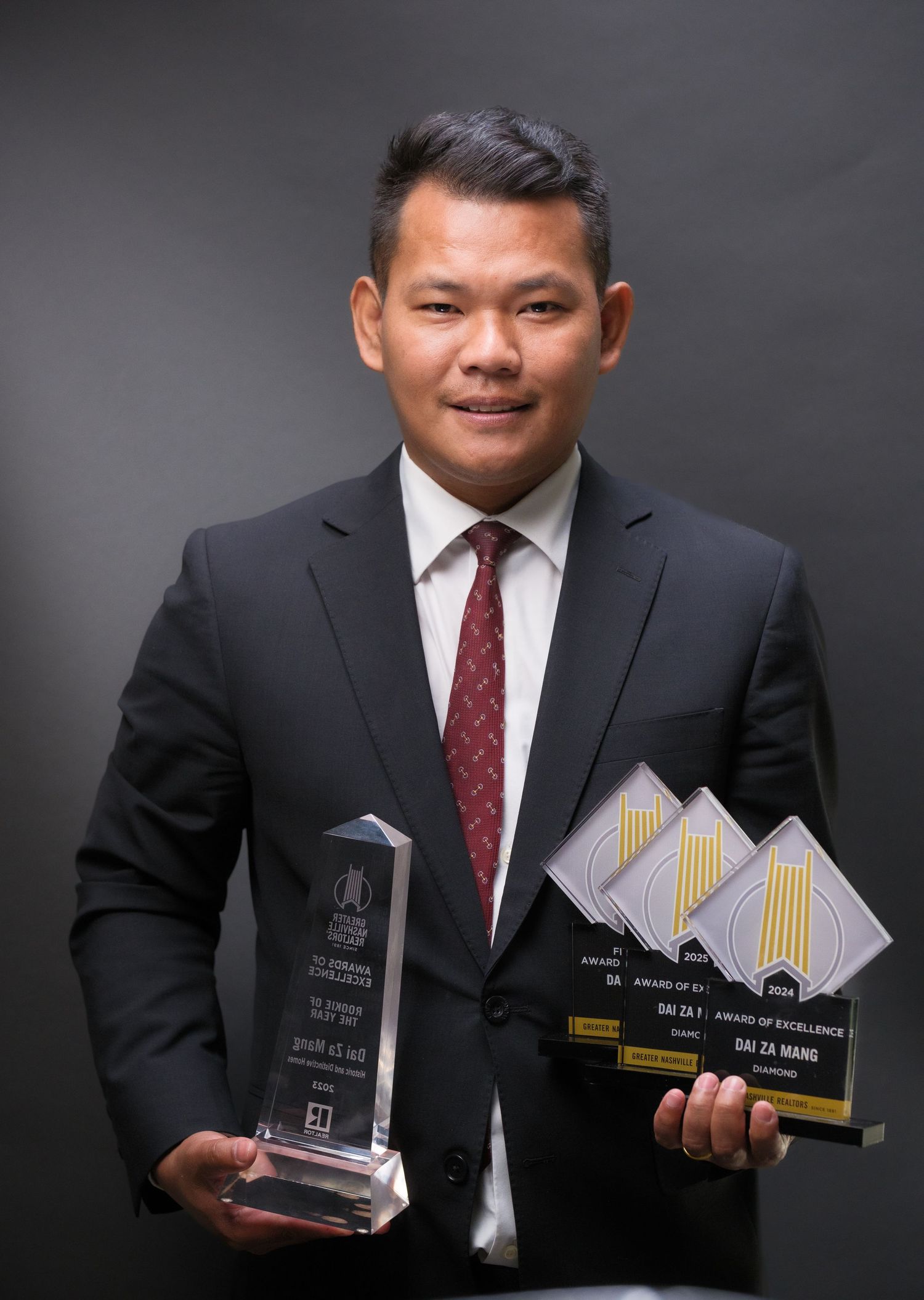 Man in suit holding crystal trophy and Award of Excellence plaques, posing proudly for professional photo.