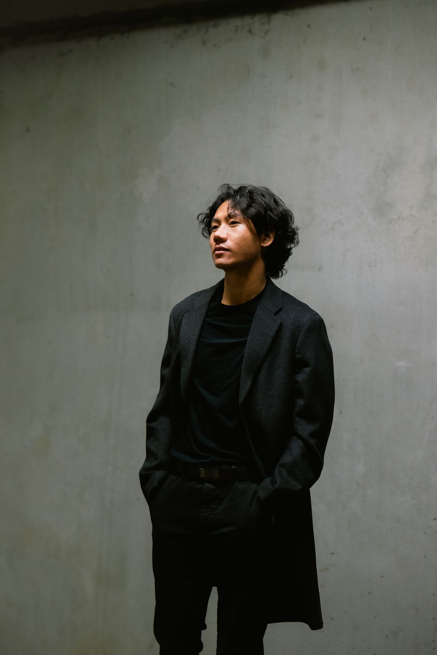 Stylish portrait against concrete wall featuring black coat and casual attire in moody lighting.