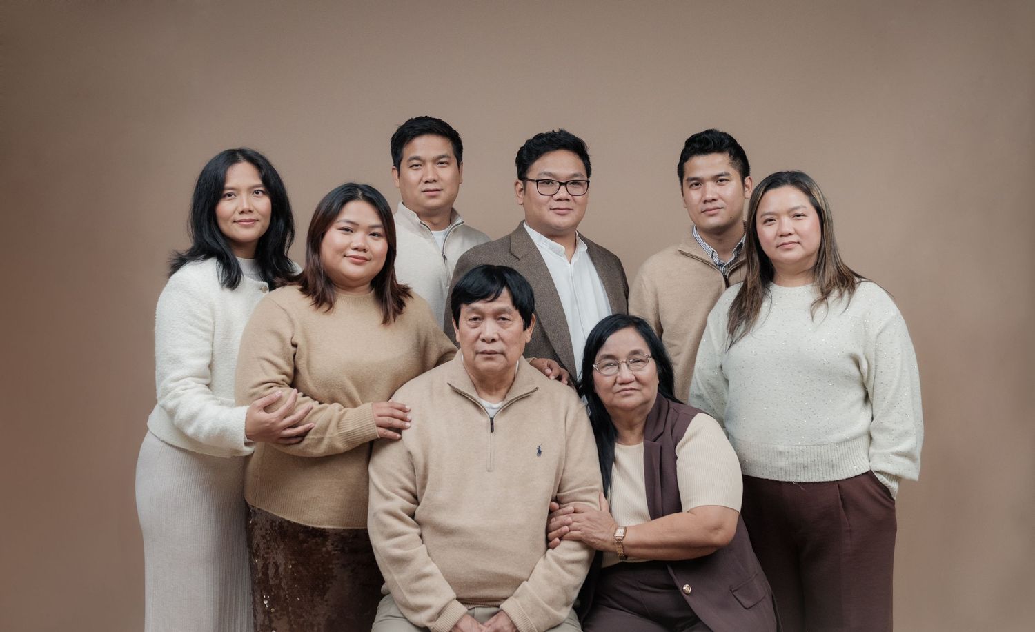 A family portrait of eight people in neutral-toned clothing against a beige background.
