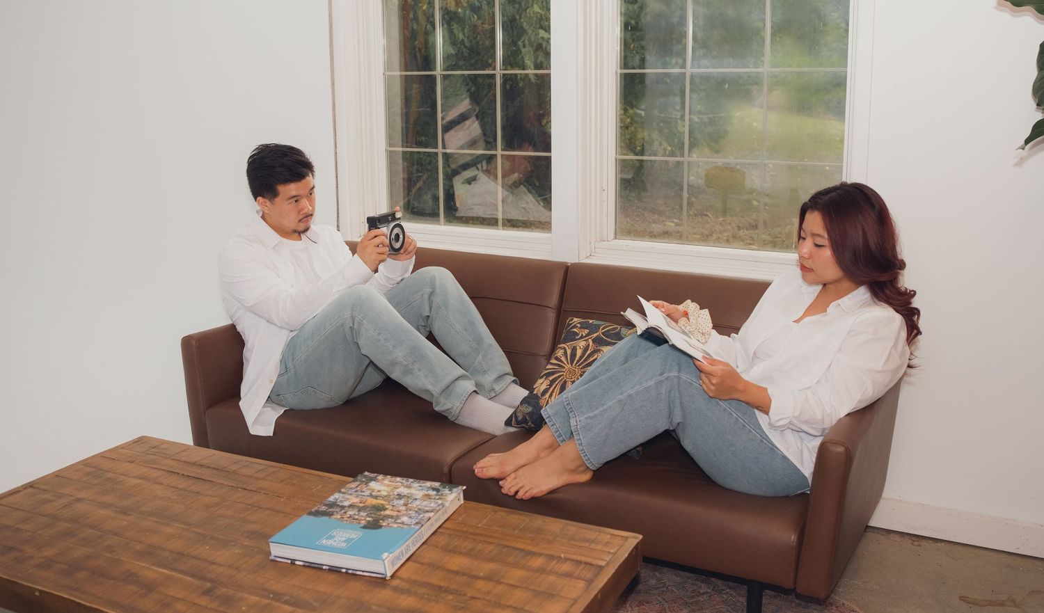 Two people in matching outfits share playful moments on a leather couch by window.