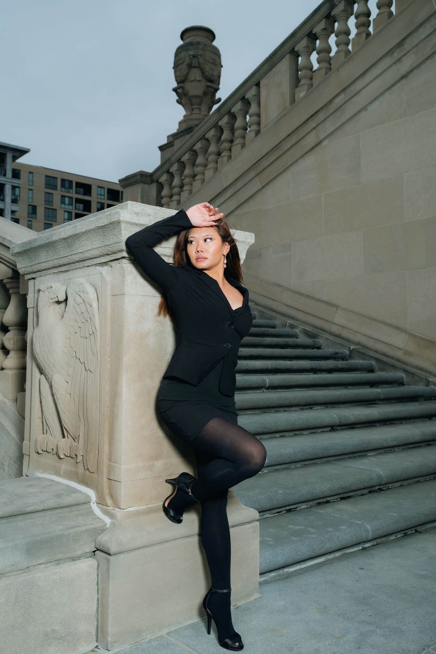 Stylish figure strikes dramatic pose against elegant stone staircase in urban setting.