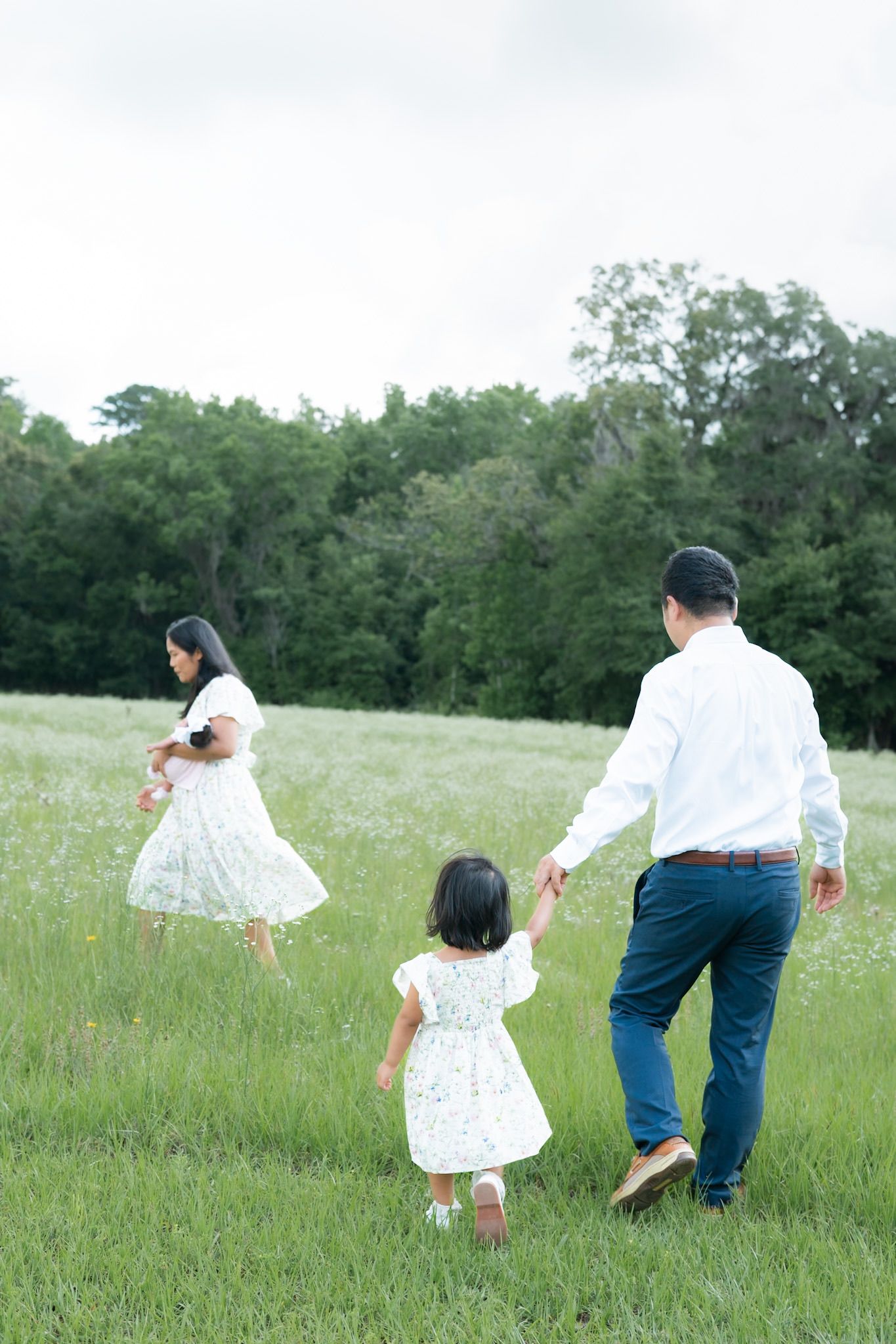 Family walking in green meadow, parents and young daughter in white outfits holding hands near lush trees.