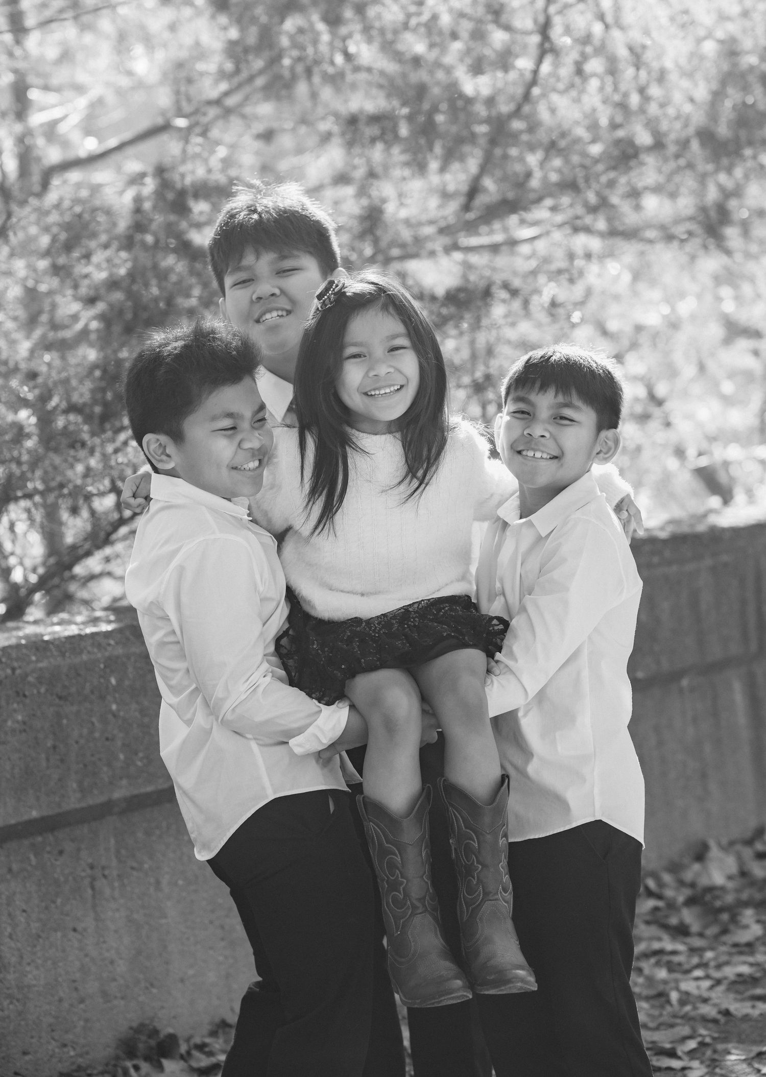 Four children pose together outdoors in a black and white photo, with three boys holding a smiling girl in cowboy boots.