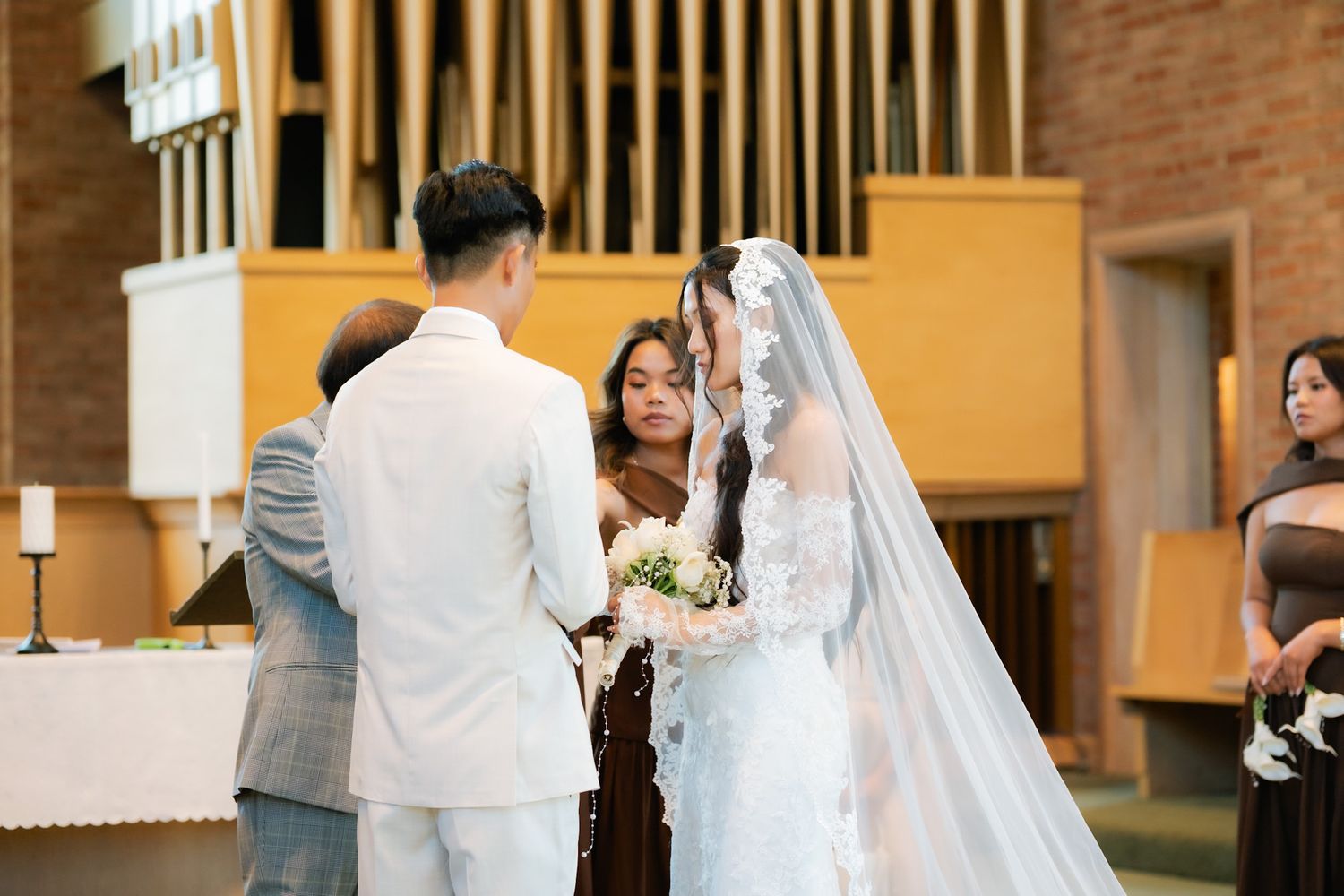 Asian couple exchanging vows at a church wedding ceremony, bride in lace gown and veil holding bouquet.
