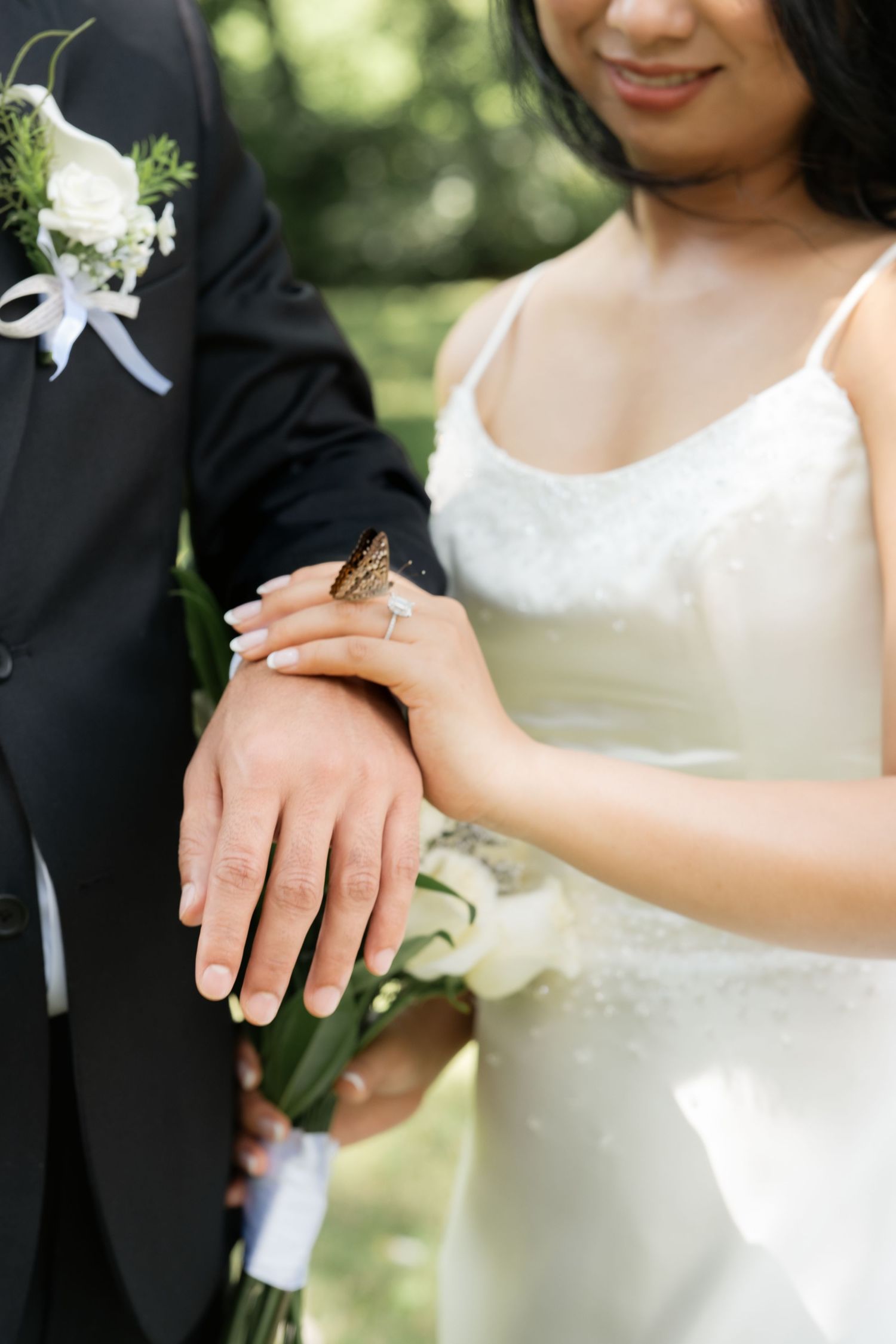 Bride and groom holding hands showing wedding rings with a butterfly perched on their hands during outdoor ceremony.