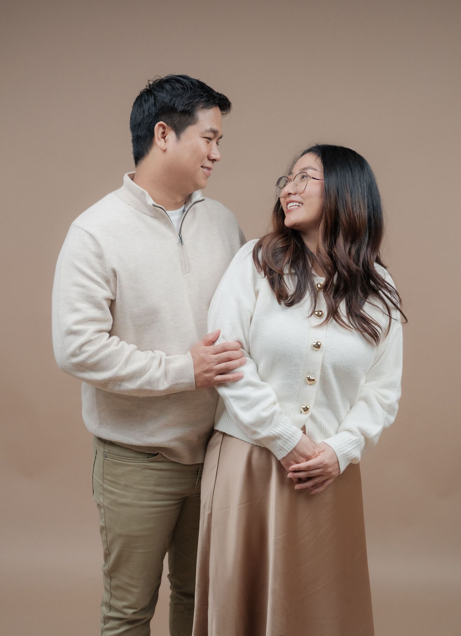 Couple in matching cream sweaters share joyful moment against brown background.
