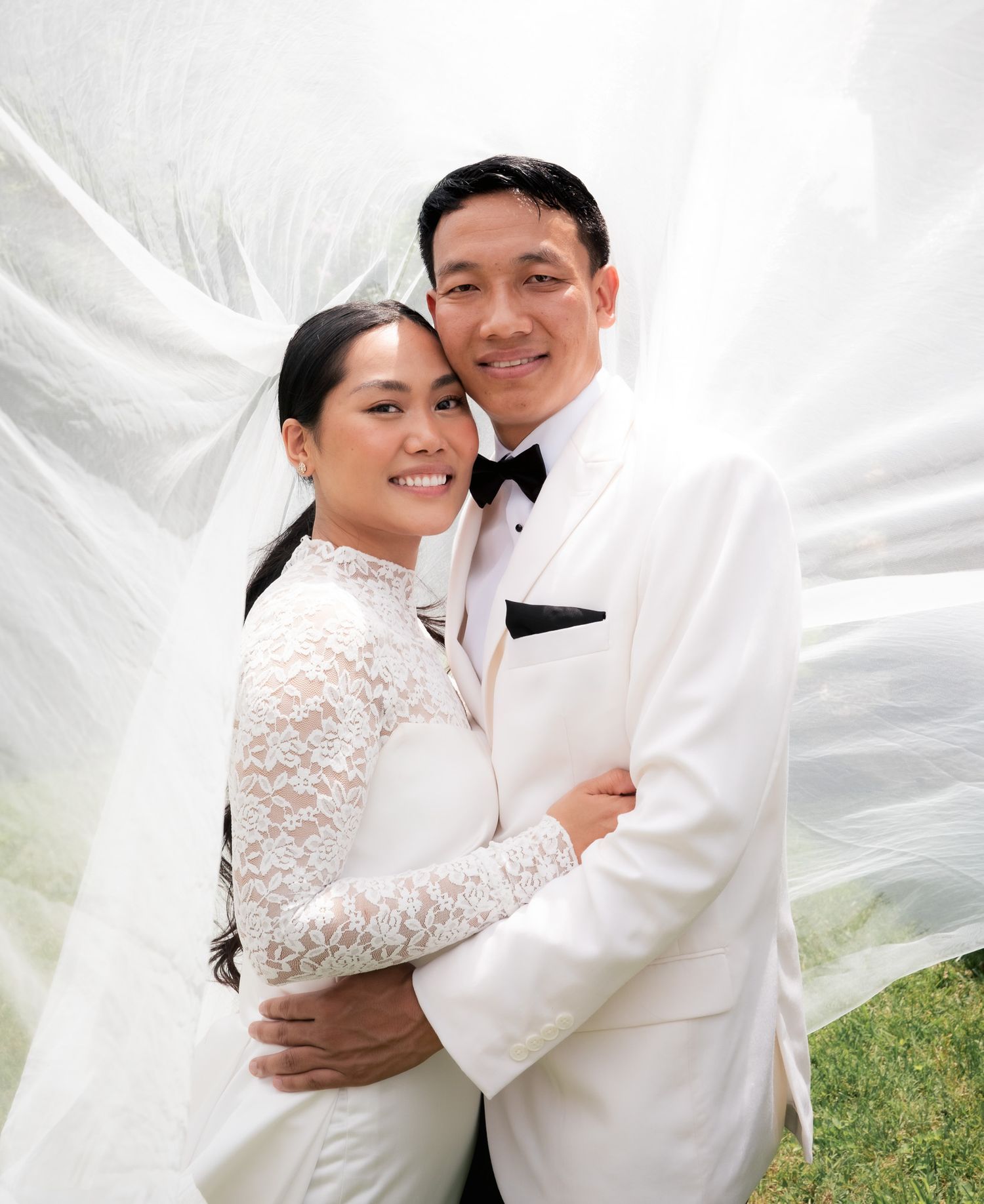 Wedding couple embracing under flowing white veil in outdoor garden setting.