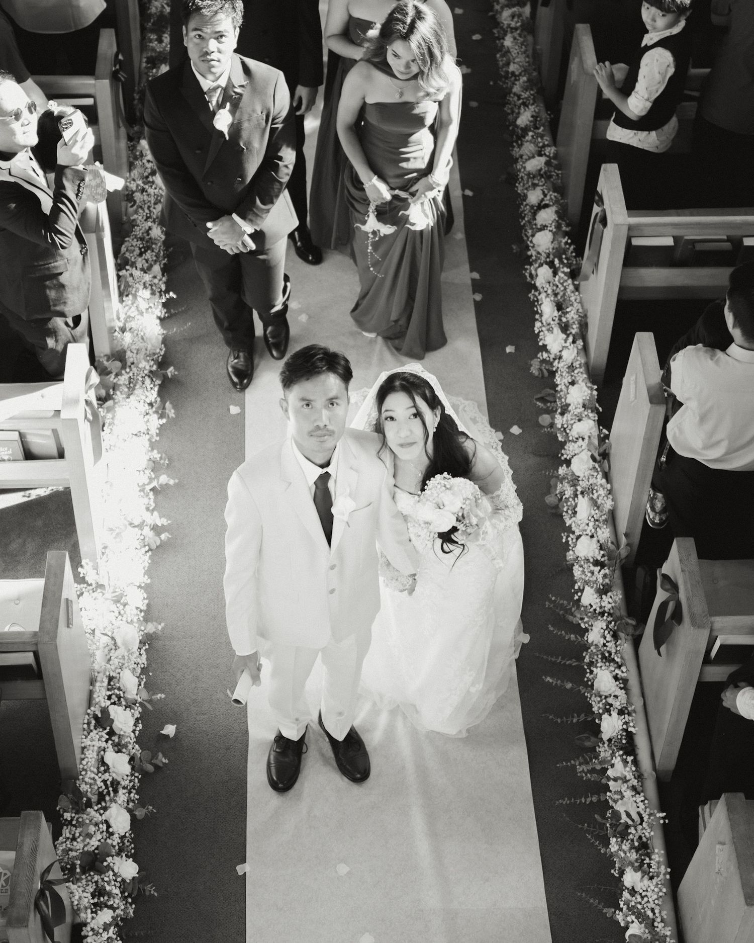 Black and white aerial view of bride and groom walking down flower-lined church aisle with wedding guests seated in pews.