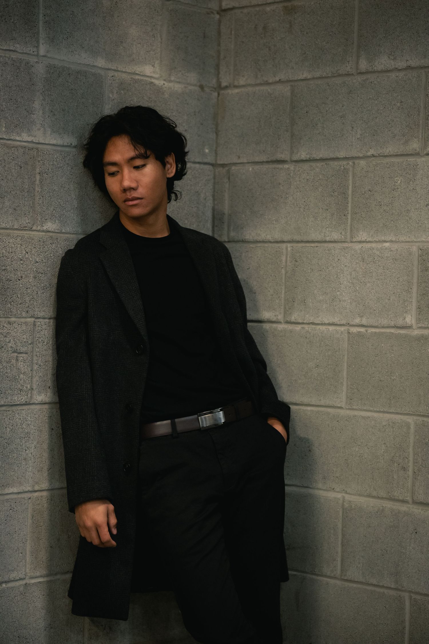 Figure in all black attire poses against a concrete wall in dramatic lighting.