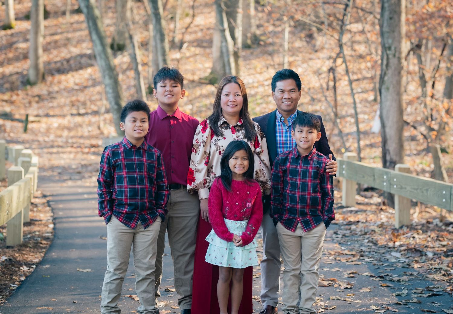 Family of six posing on a wooden bridge path surrounded by autumn trees in a park during fall season.