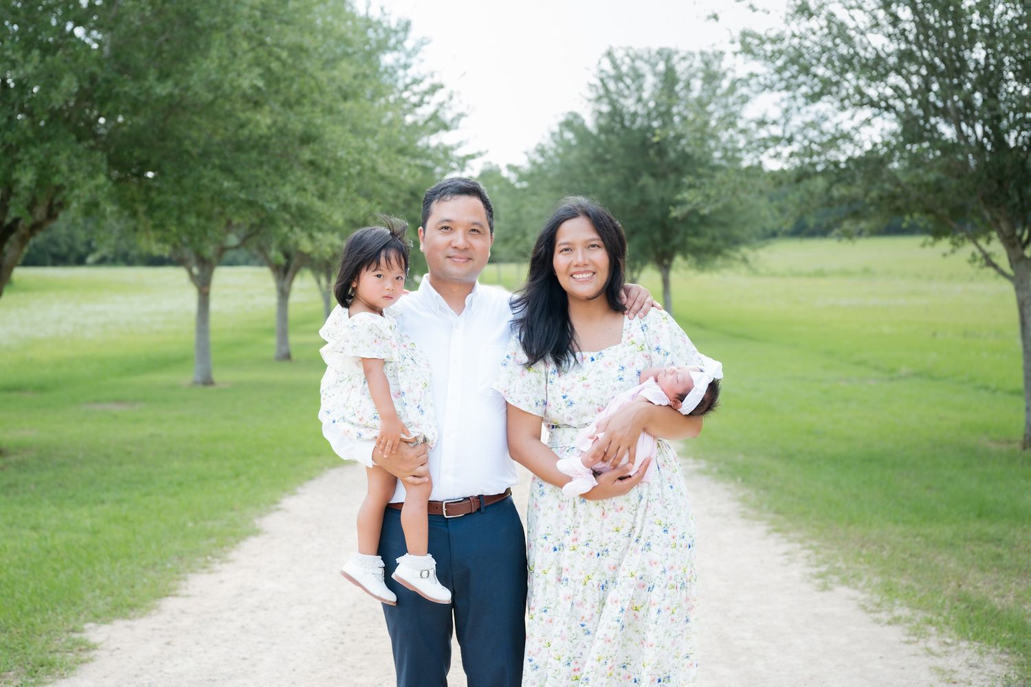 Family of three poses on a park path, parents holding two young children among lush green trees.