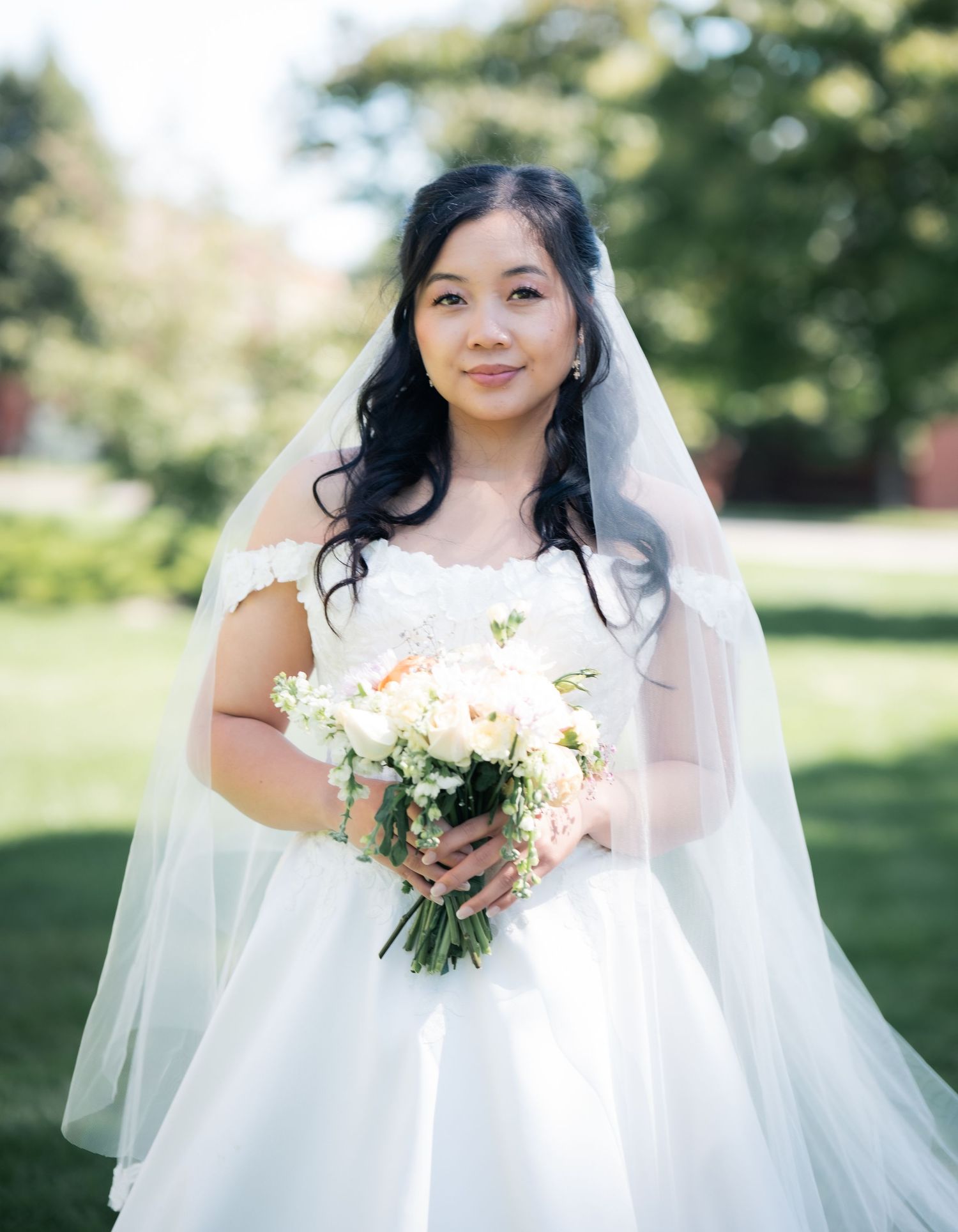 Bride in white wedding gown holds bouquet while posing in outdoor garden setting.