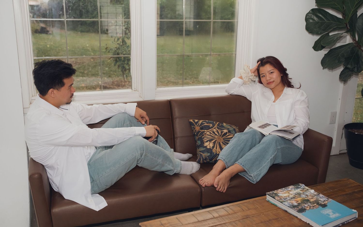 Playful couple lounges on brown leather sofa in bright room with large windows.