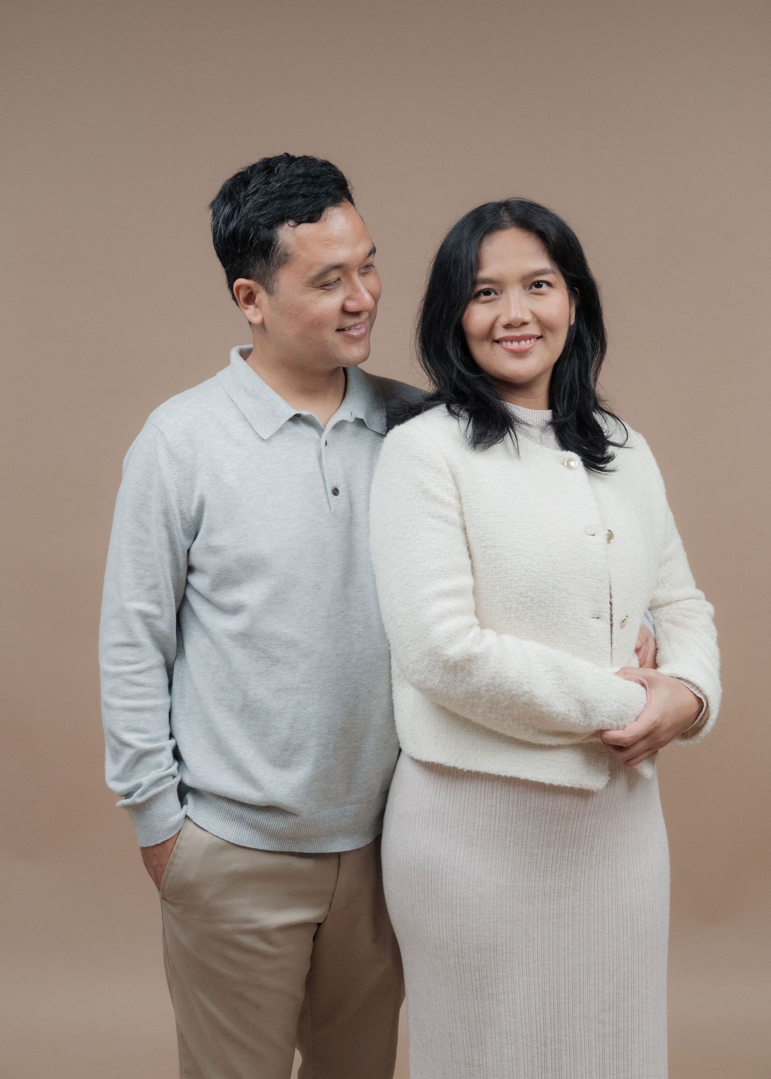 A couple poses together for maternity photos wearing coordinated light-colored outfits against a neutral backdrop.