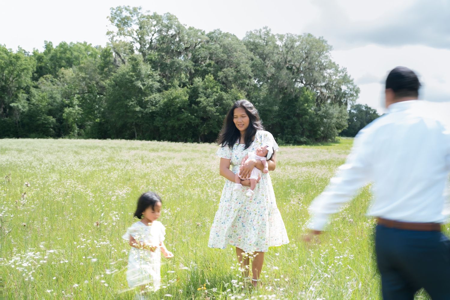 Mother in white dress holding newborn with toddler beside her in a sunny wildflower meadow.