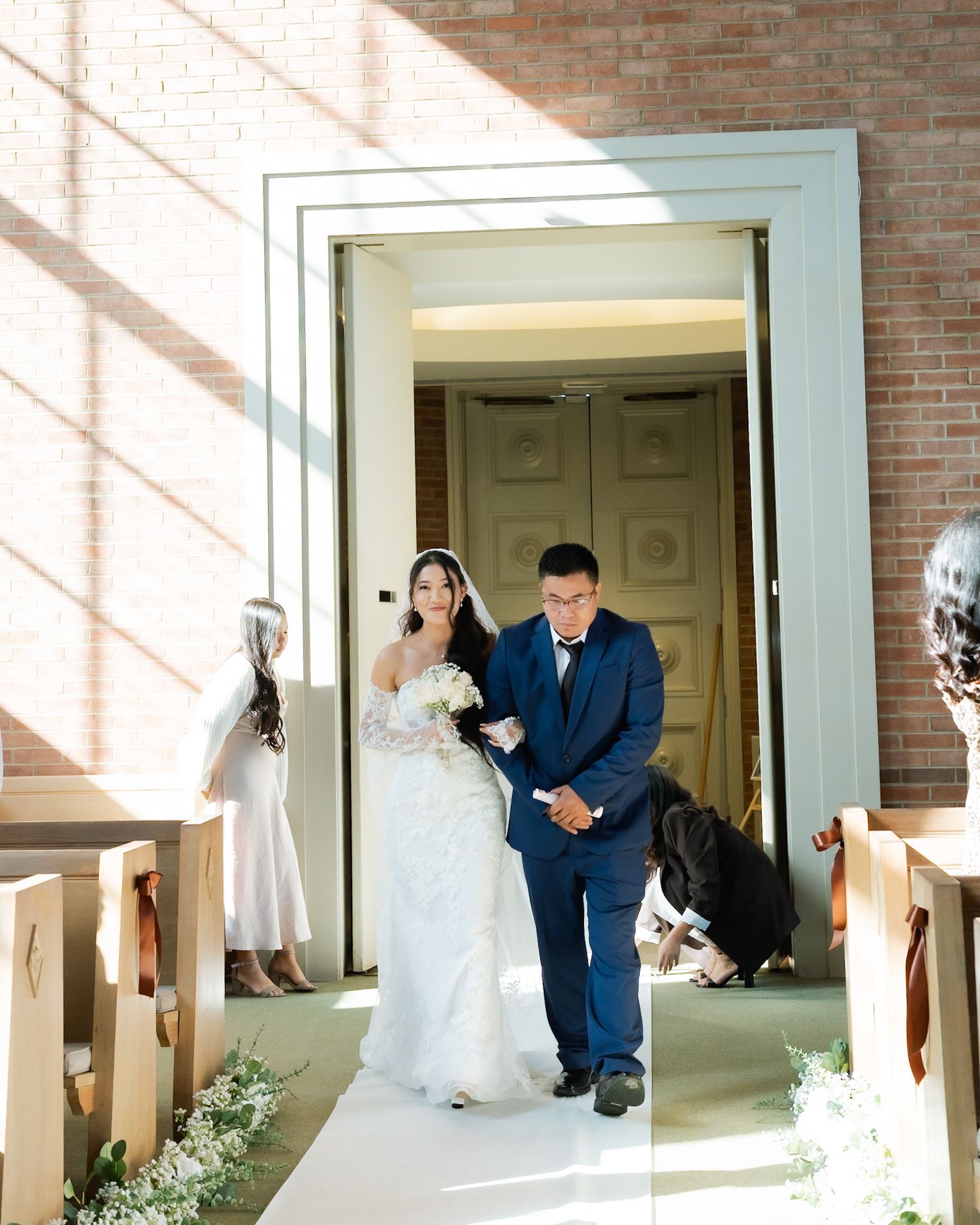 Bride in white gown walks down church aisle with escort in navy suit during elegant wedding ceremony.