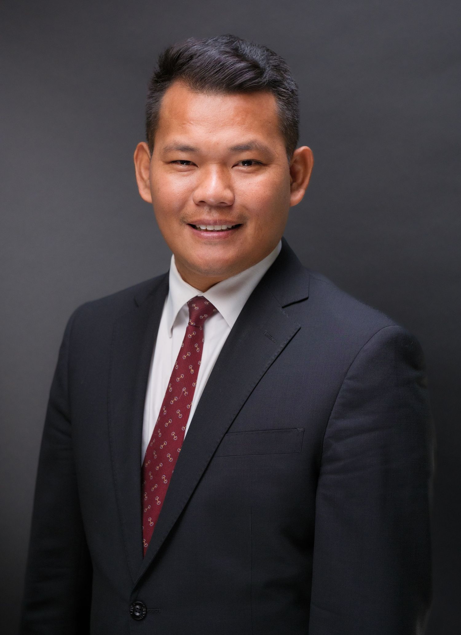 Professional headshot of a smiling Asian man in a dark suit and red tie against a gray background.