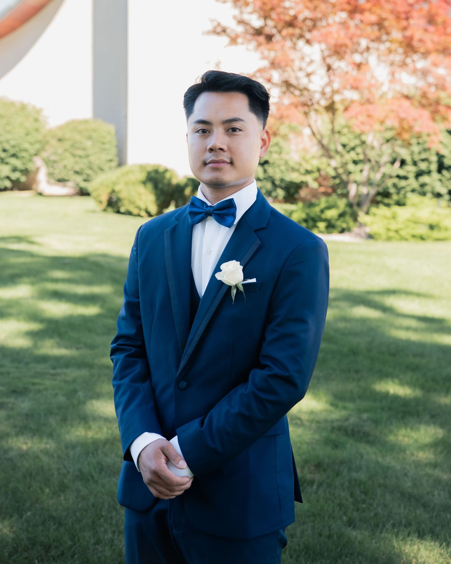 Groom in navy tuxedo poses in garden setting with autumn foliage.