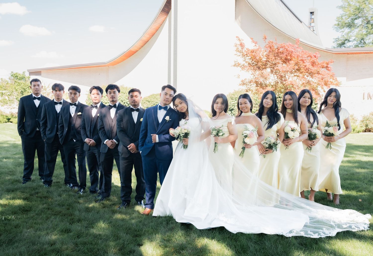 Wedding party members in navy suits and champagne dresses pose together on a sunny lawn.