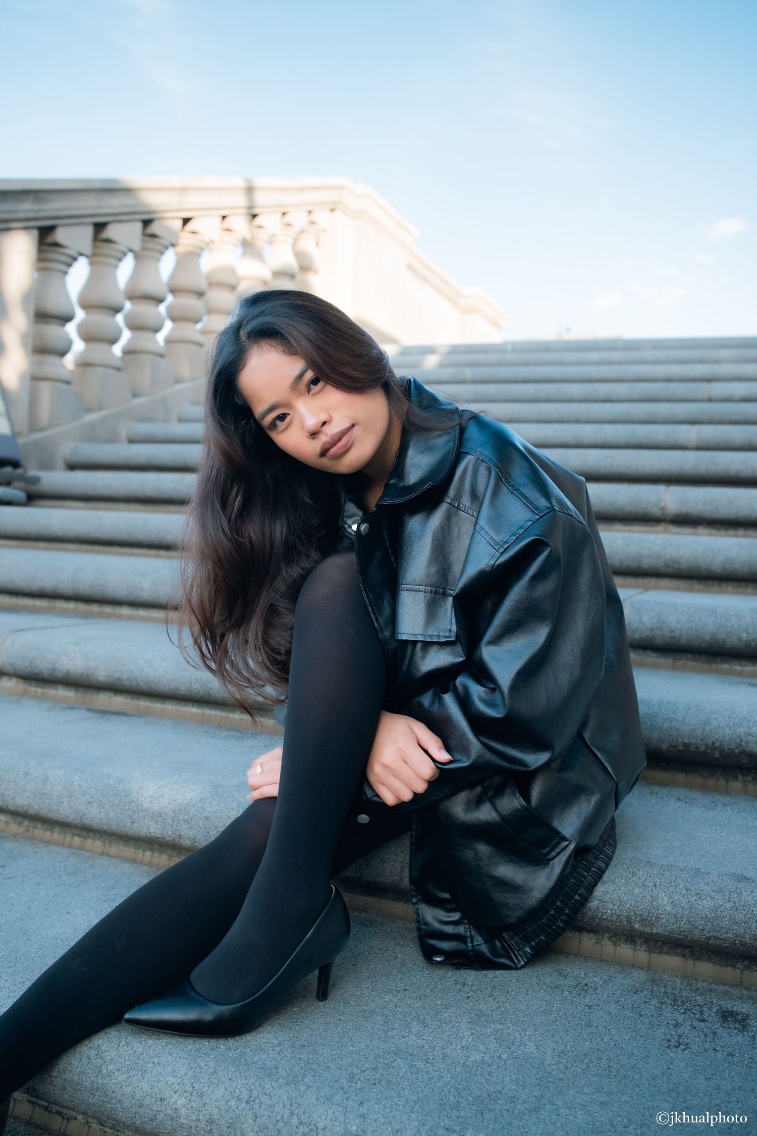 A person in a black leather jacket poses on concrete steps during a moody outdoor photoshoot.