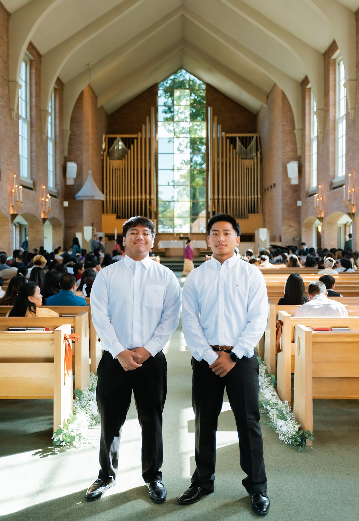 Two young men in white shirts and black pants stand in a sunlit church aisle during a wedding ceremony.