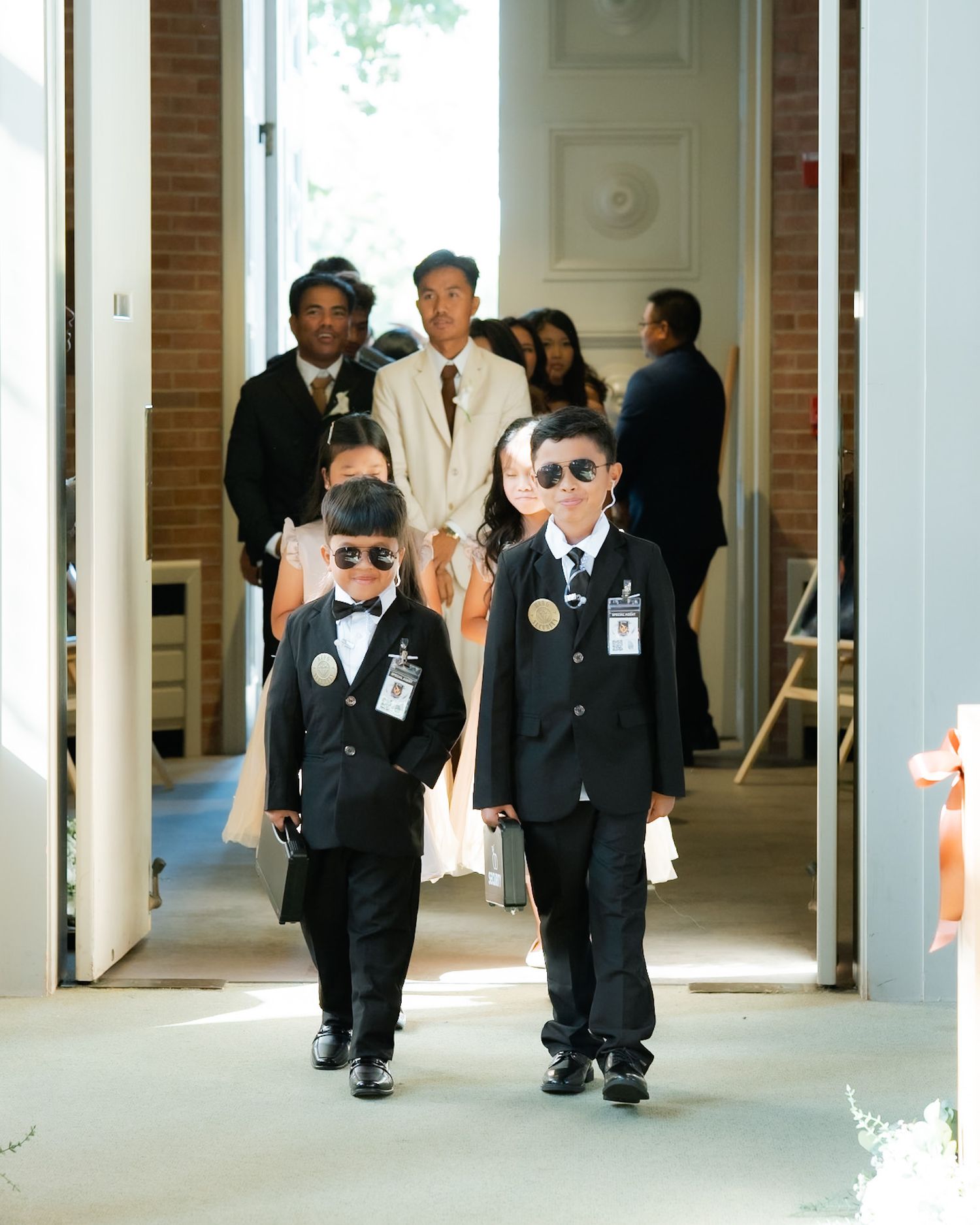 Two young boys in black tuxedos and sunglasses walk down a wedding aisle, styled as mini secret agents.